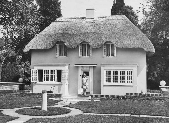 Cottage with a thatched roof and large windows. Woman standing at the entrance holding a small bag. Garden with pathway and trees