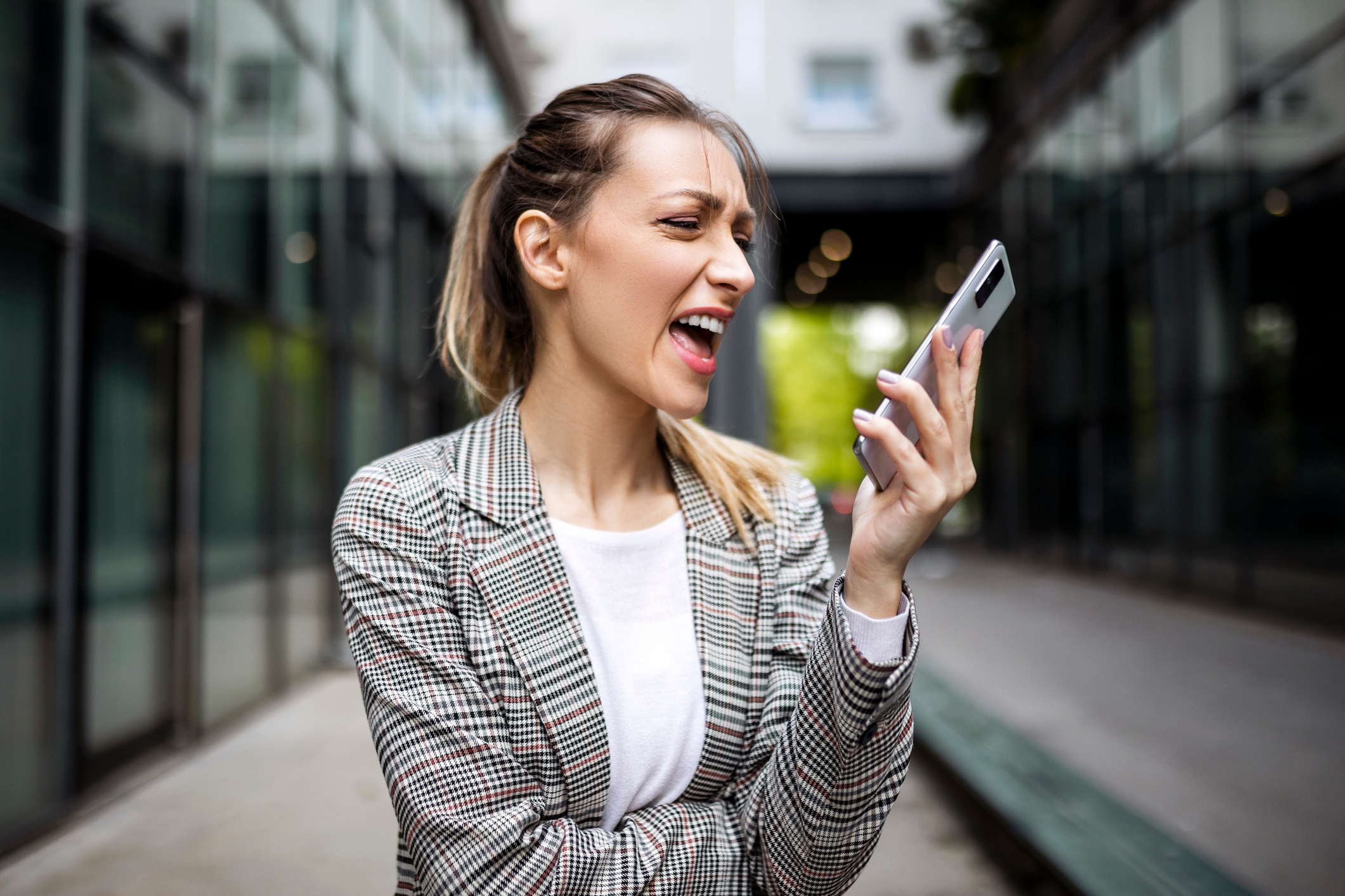 A woman in a plaid blazer excitedly shouts at her phone while standing outside between glass buildings