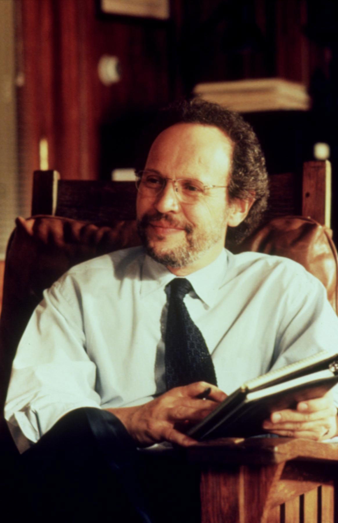 Billy Crystal seated, wearing a shirt and tie, holding a book with a thoughtful expression in a cozy setting