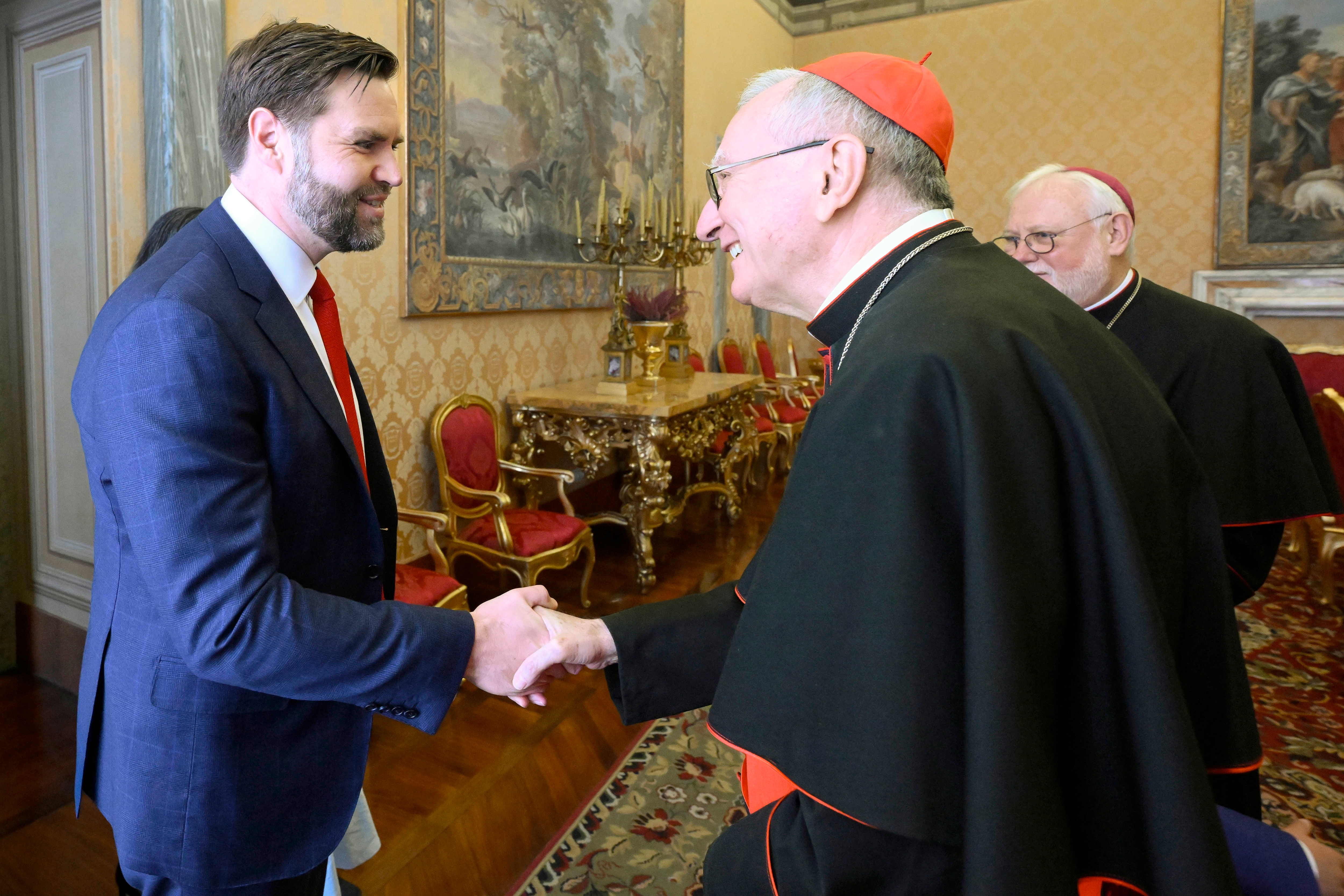 Two people shaking hands in a formal, ornate room. One wears religious attire with a red cap; both appear engaged in a polite exchange