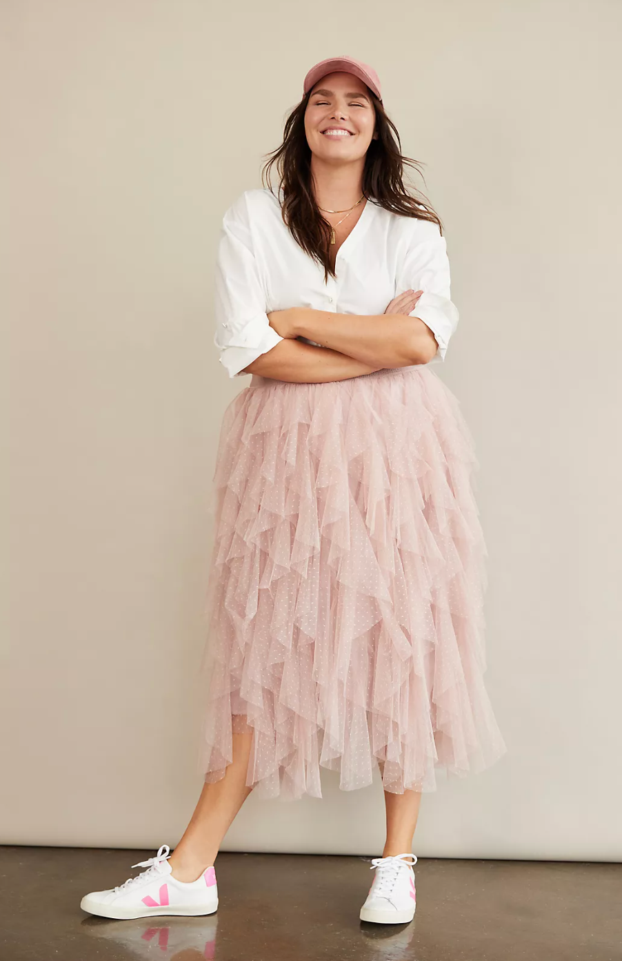 Woman smiling, wearing a white blouse, pink tulle skirt, pink cap, and white sneakers with pink details, posing against a plain background