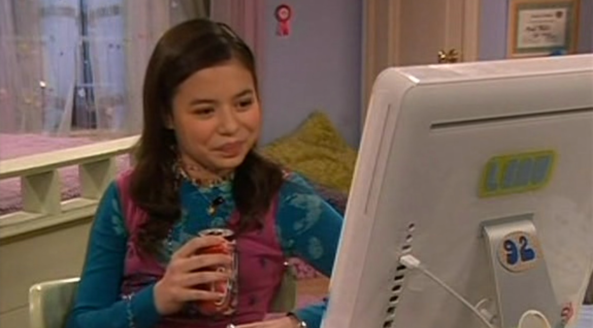 Miranda Cosgrove sits at a computer in a bedroom, holding a can, and smiling at the screen