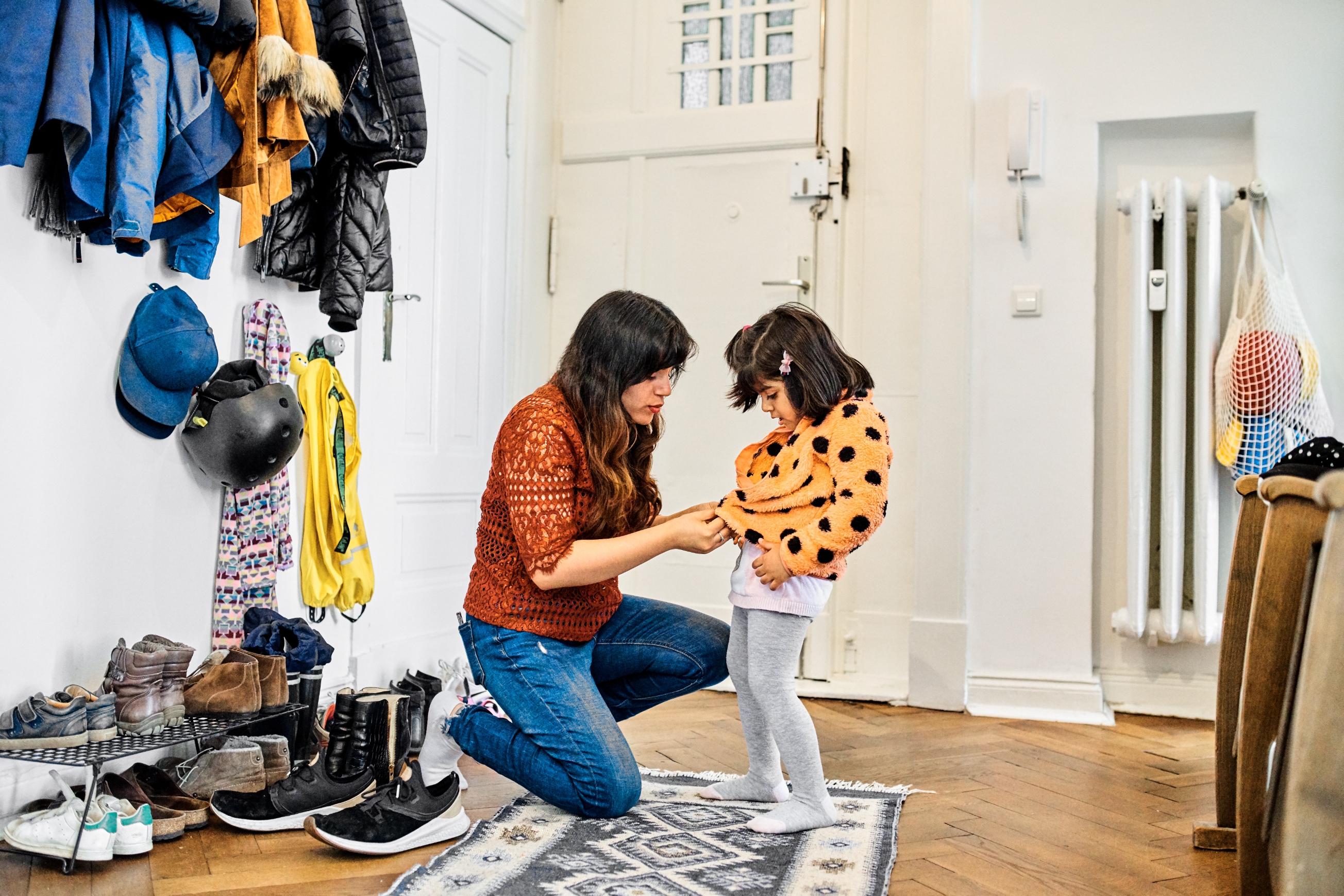 Woman kneeling, helping child with coat in an entryway lined with jackets and shoes. Cozy, organized setting