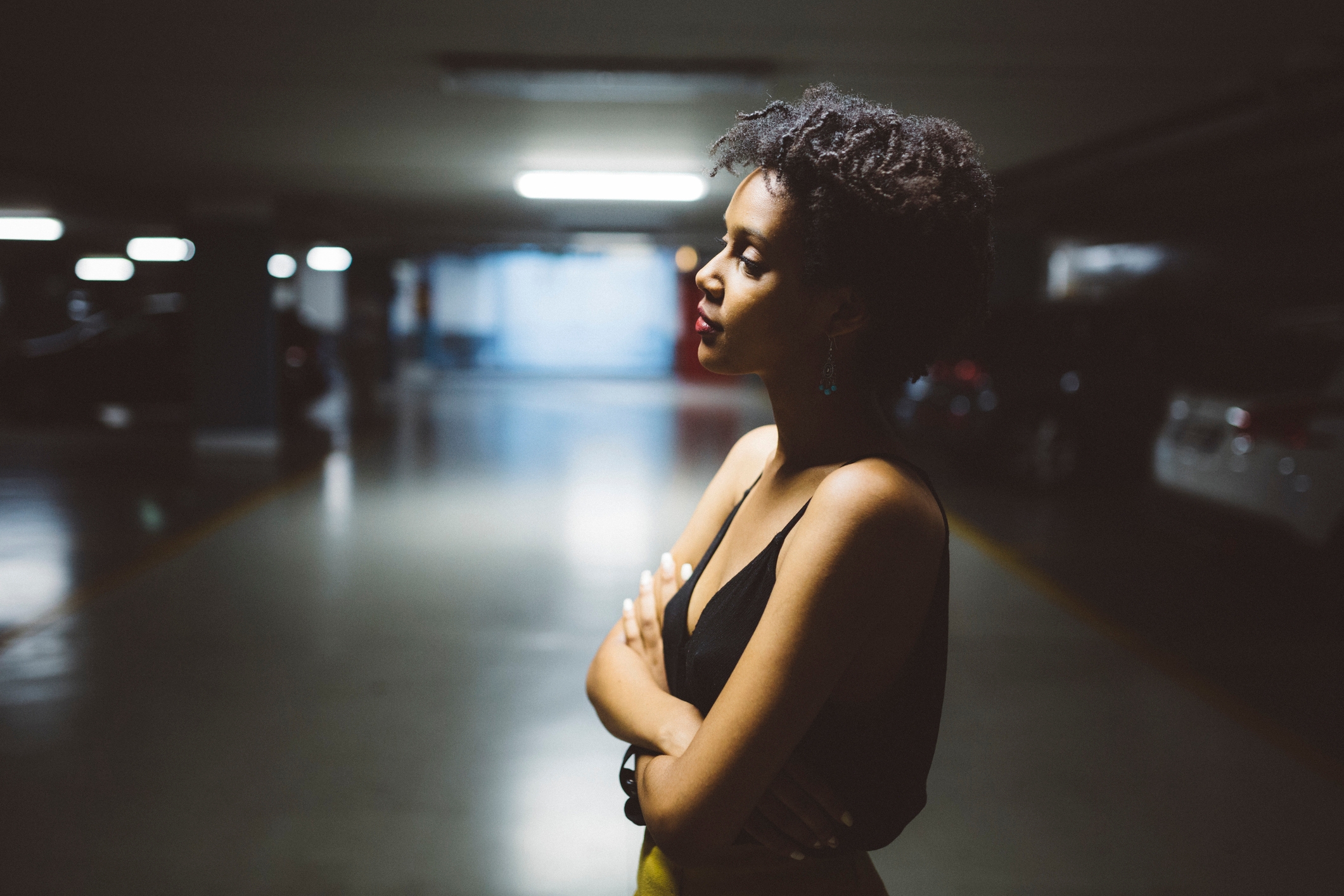 Person in a dimly lit parking garage, standing confidently with arms crossed. They are wearing a sleeveless top and have curly hair