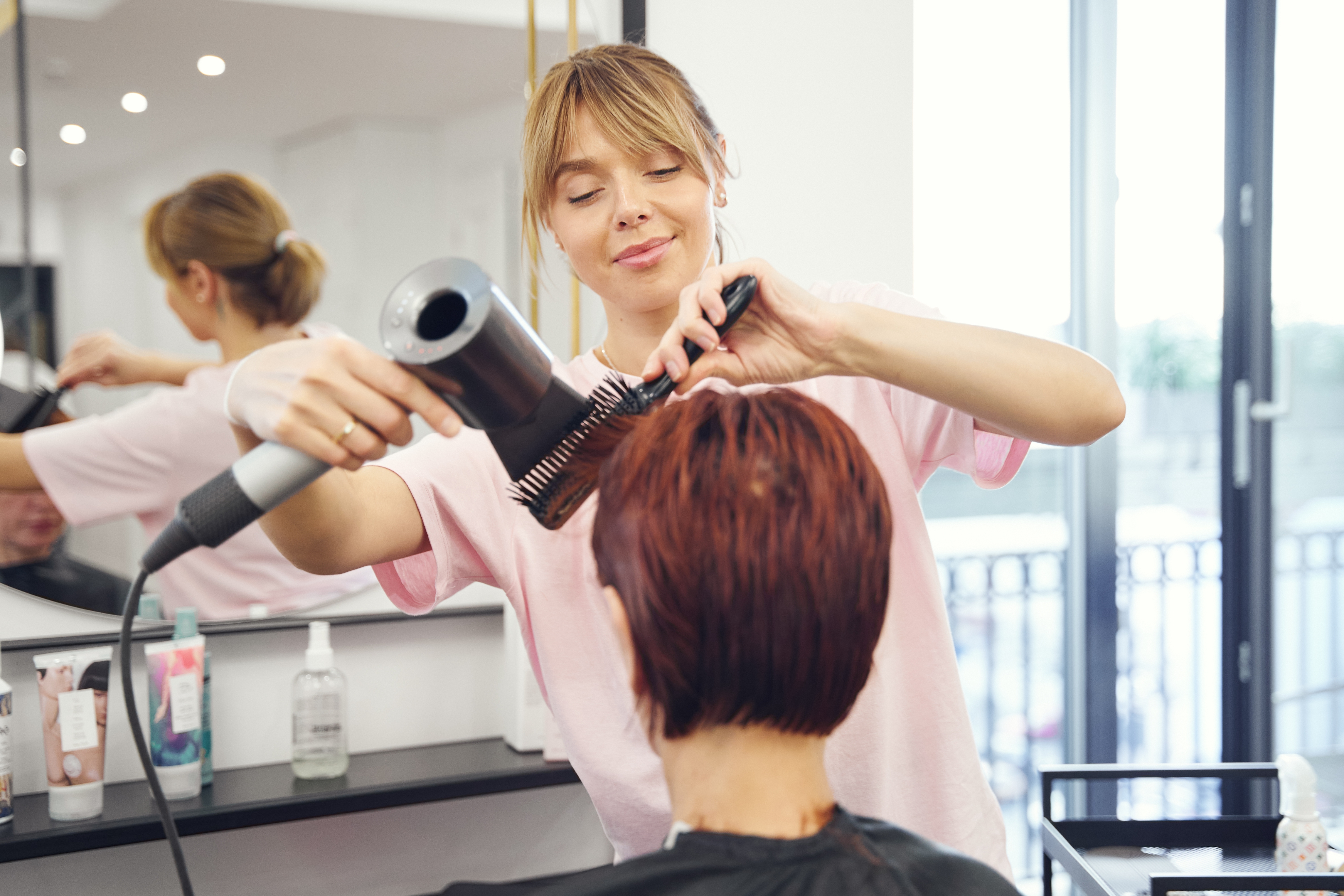 Hairdresser blow-dries and brushes short hair in a salon, focusing on a client's new hairstyle