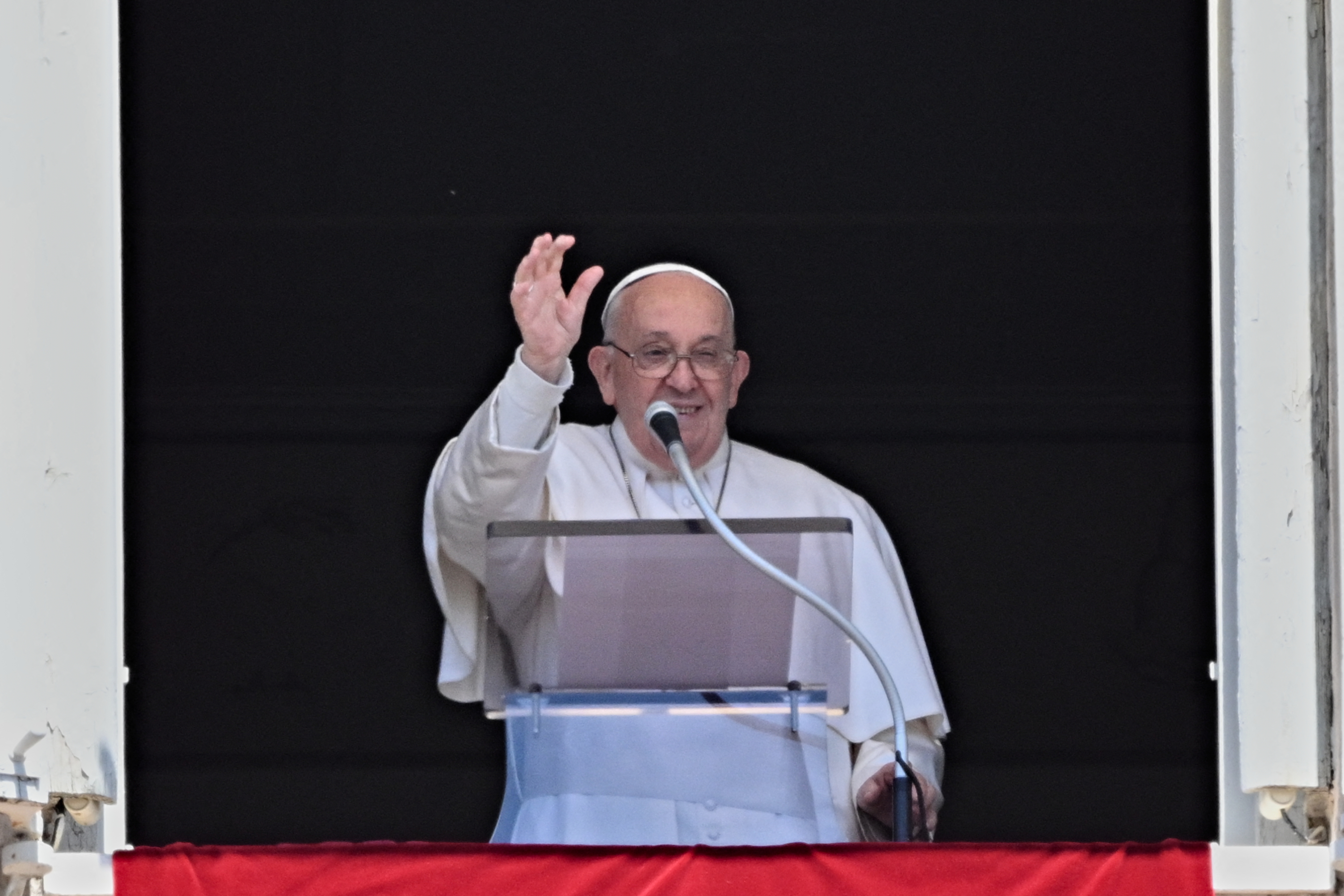 Pope Francis wearing religious attire speaks from a balcony, gesturing with one hand, behind a podium with a microphone