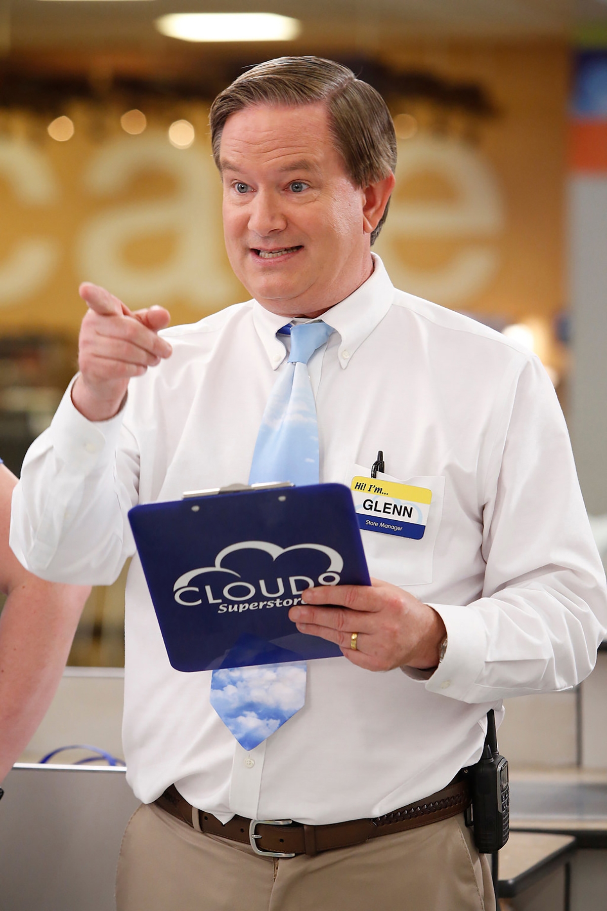 Man in a white shirt and patterned tie holding a clipboard, speaking energetically, in a store setting