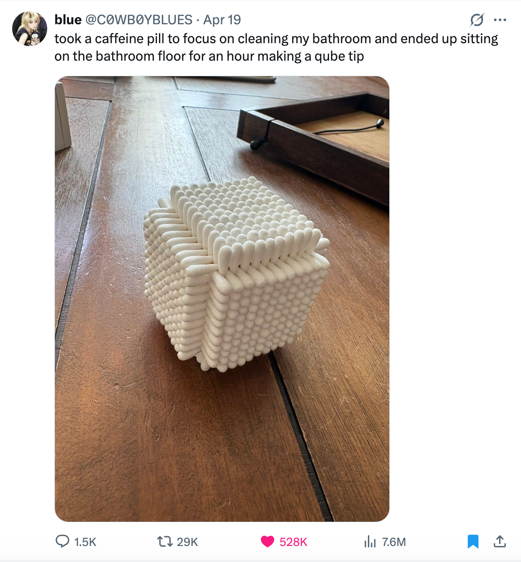 A cube made of numerous Q-tips sits on a wooden floor. A humorous tweet describes a caffeine-fueled cleaning session ending in making the cube