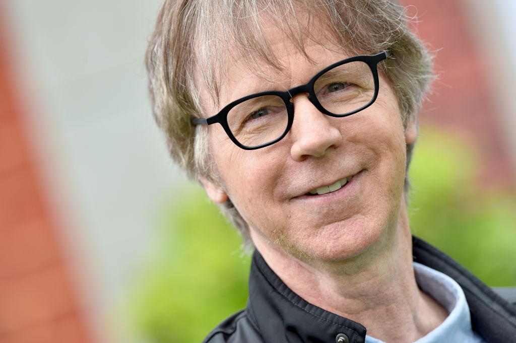A person wearing black glasses and a jacket, smiling outdoors, with blurred greenery in the background