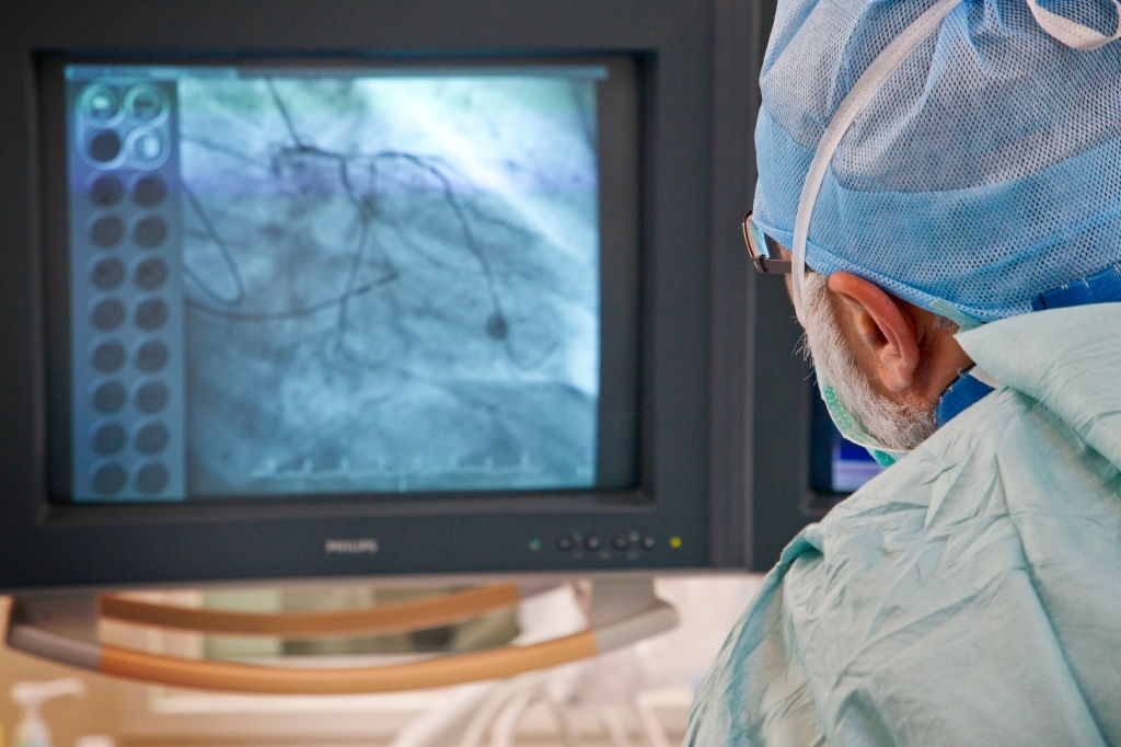 A surgeon in scrubs and a mask examines a medical scan on a monitor during a procedure