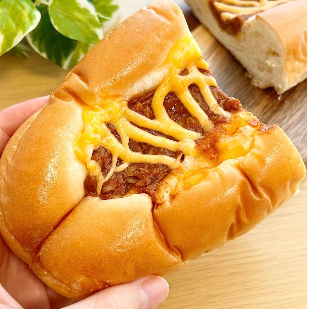 手に持たれたパンに、チーズと肉がトッピングされています。背景には切られた別のパンがあります。