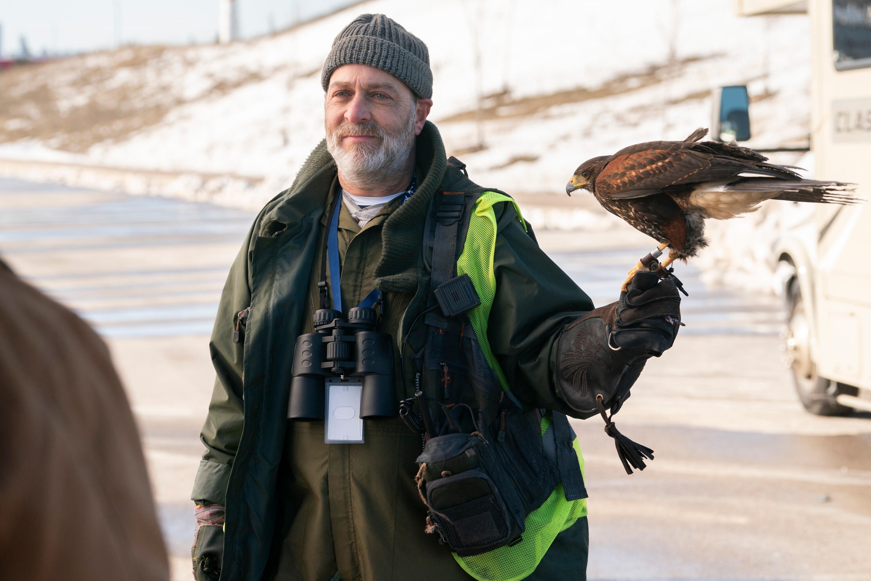 Man in outdoor gear and beanie holding a hawk, with a pair of binoculars around his neck, standing near a snowy road