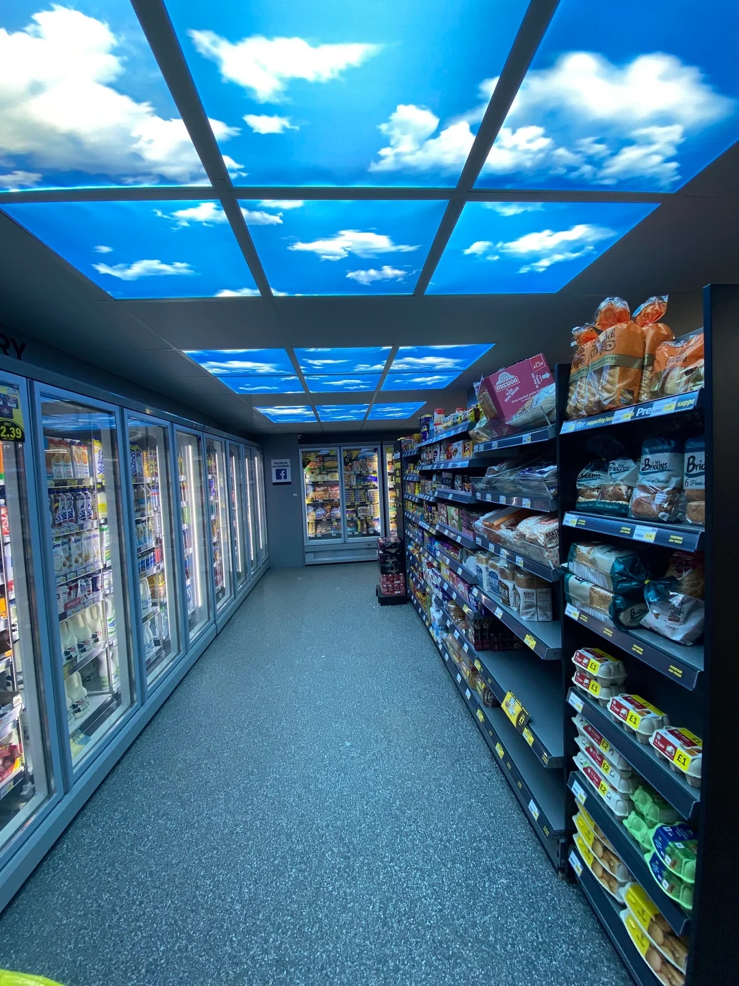 Grocery store aisle with fridge and shelves, featuring a ceiling display of blue sky with clouds