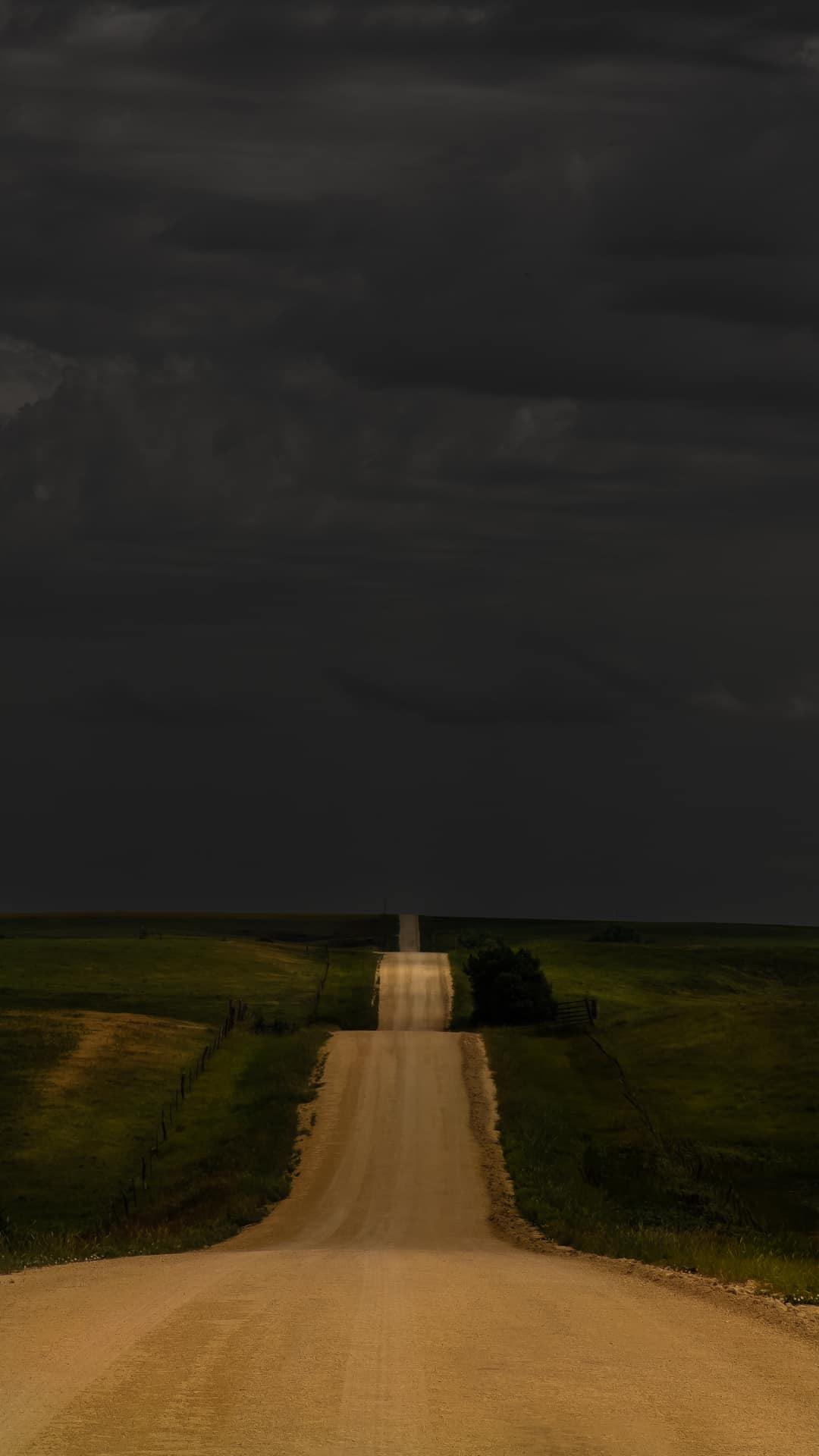A long, straight dirt road stretches into the horizon under a dark, cloudy sky, flanked by fields