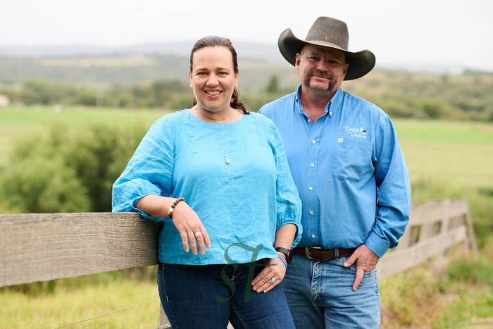 Two people smiling in a rural setting, both wearing casual blue shirts and jeans; one wears a cowboy hat. They lean against a wooden fence