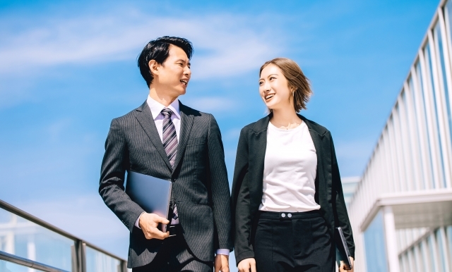 スーツ姿の男女が並んで歩きながら笑顔で会話している。背景に青空と建物が見える。