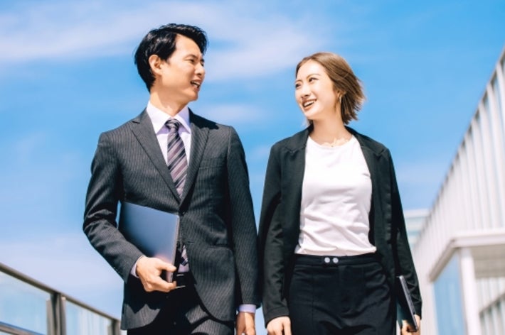 ビジネススーツを着た男女が歩きながら笑顔で会話している。青空の下でのプロフェッショナルなシーン。