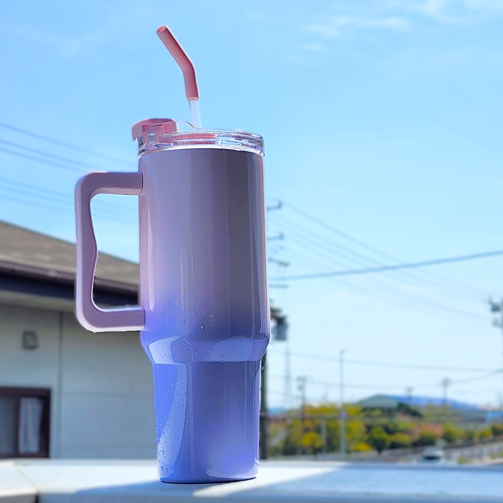 大きなハンドルとストロー付きの飲み物が入ったタンブラーが屋外に置かれている。背景に青空と建物。