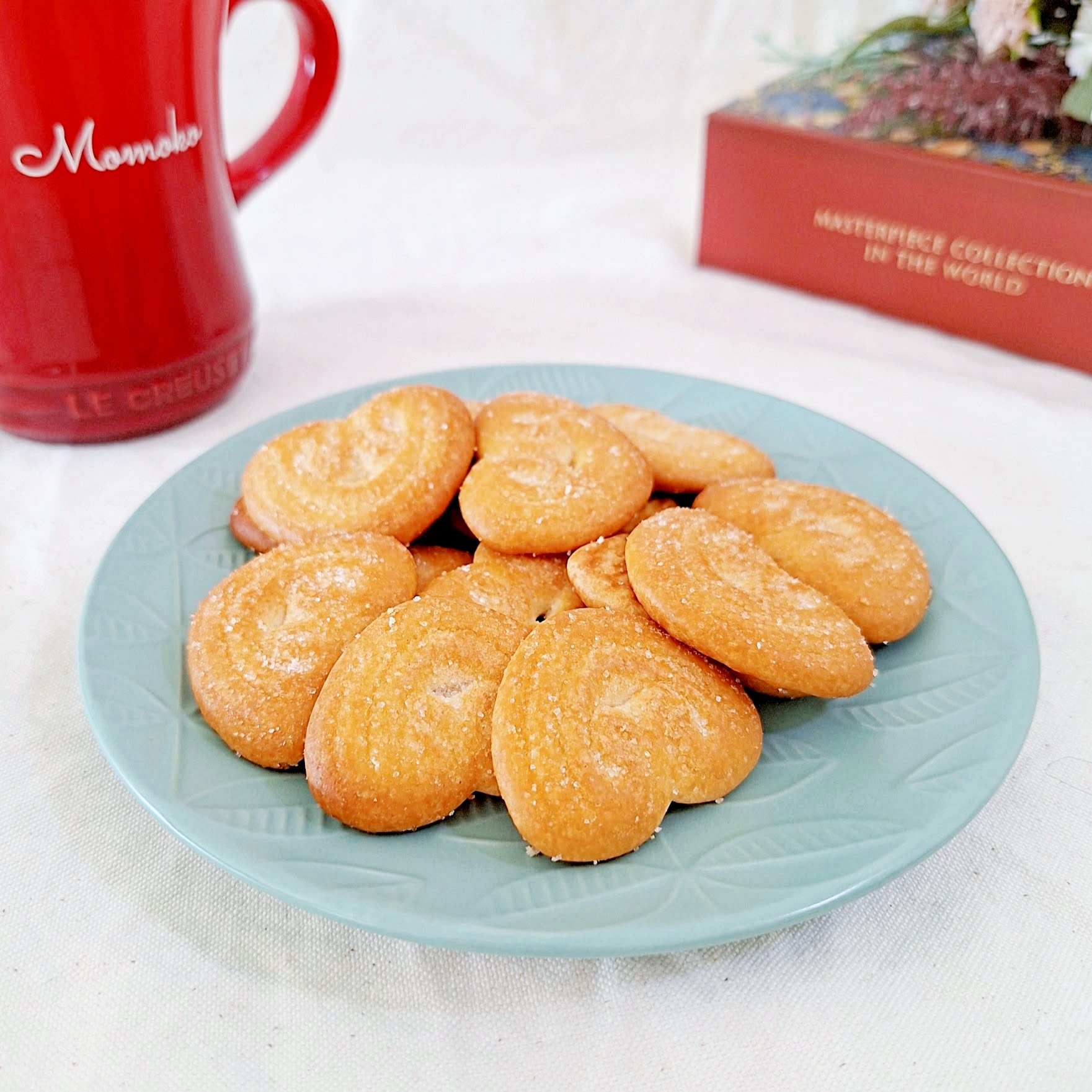 青い皿にハート形のビスケットが載っている。背景には赤いマグカップと赤い箱がある。