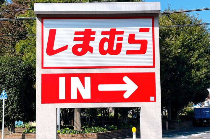 Sign with red text and arrow pointing right, indicating the direction to the store Shimamura. Trees and road in the background