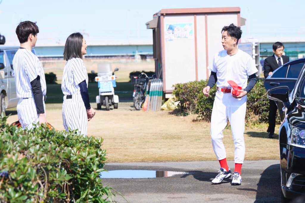 野球のユニフォームを着た3人のグループが外で会話をしている。背景には芝生と車。