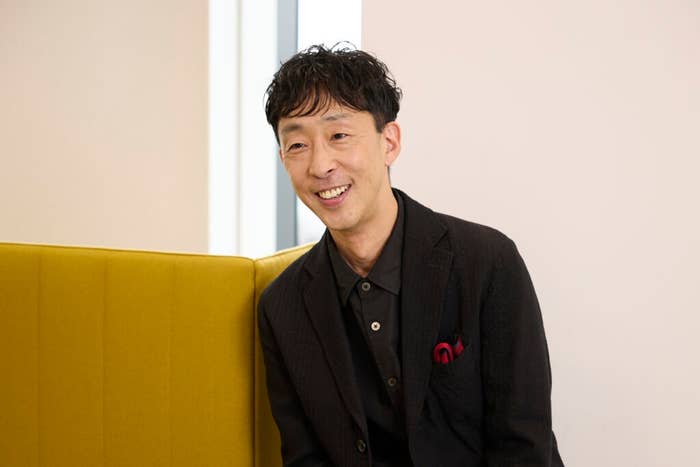 有名人と思われる男性が、シンプルな黒のスーツを着て座り、こちらを見て微笑んでいます。