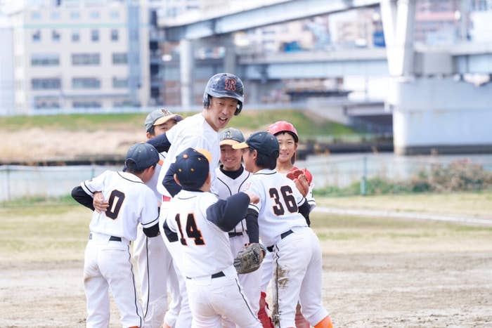 野球のユニフォームを着た子供たちのチームがグラウンドで喜び合っている。橋が背景にある。