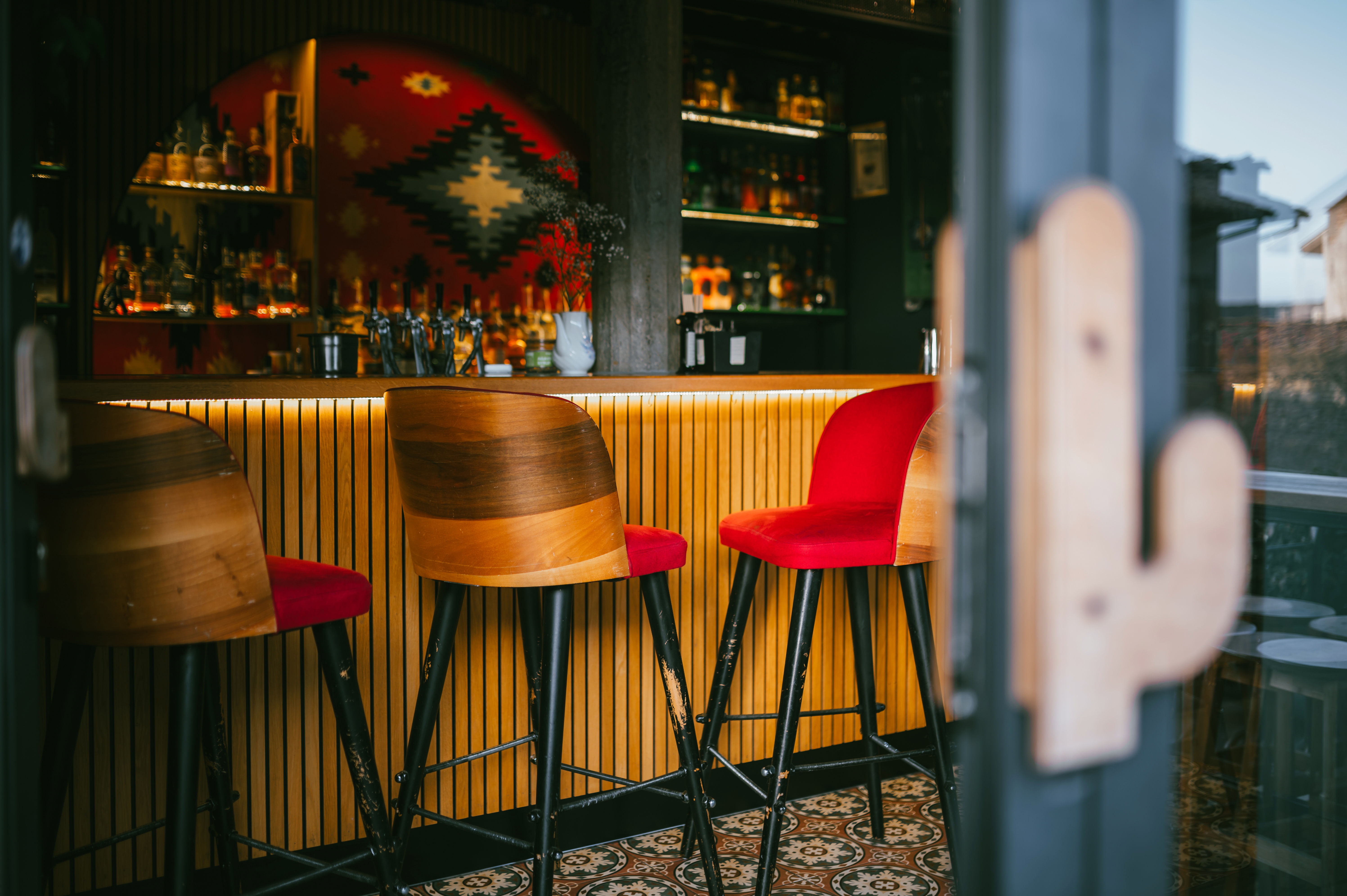 Bar interior with wooden and red chairs, a patterned tile floor, and a backlit bar counter with various bottles displayed