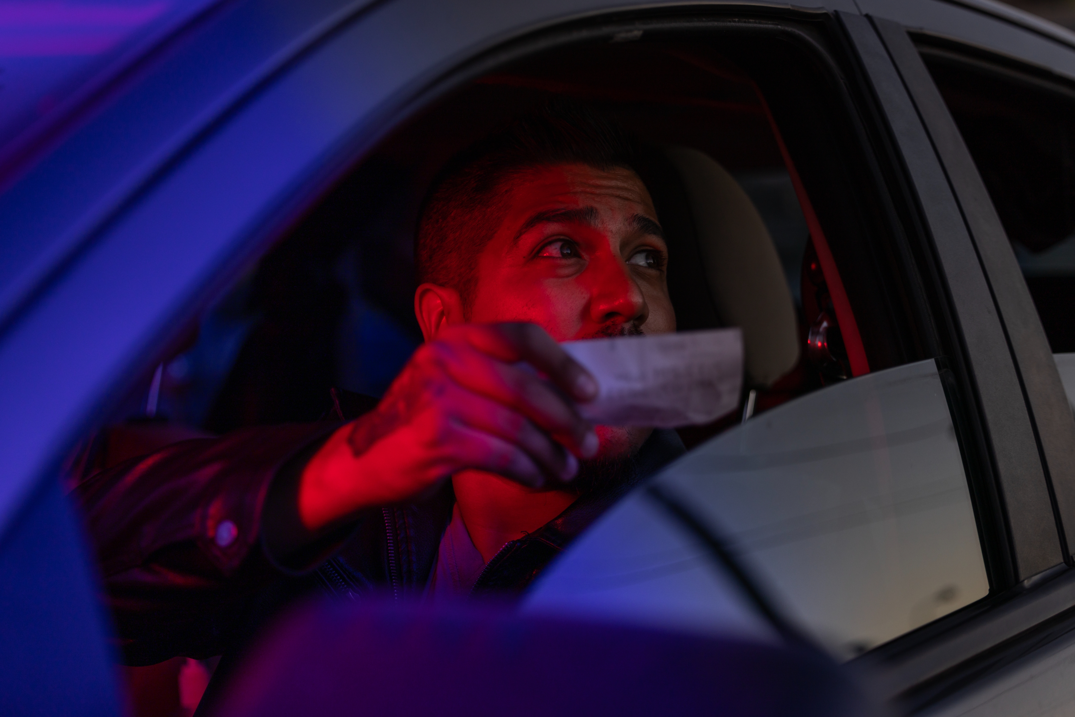 Person in a car showing a document to someone outside, illuminated by intense lighting through the window