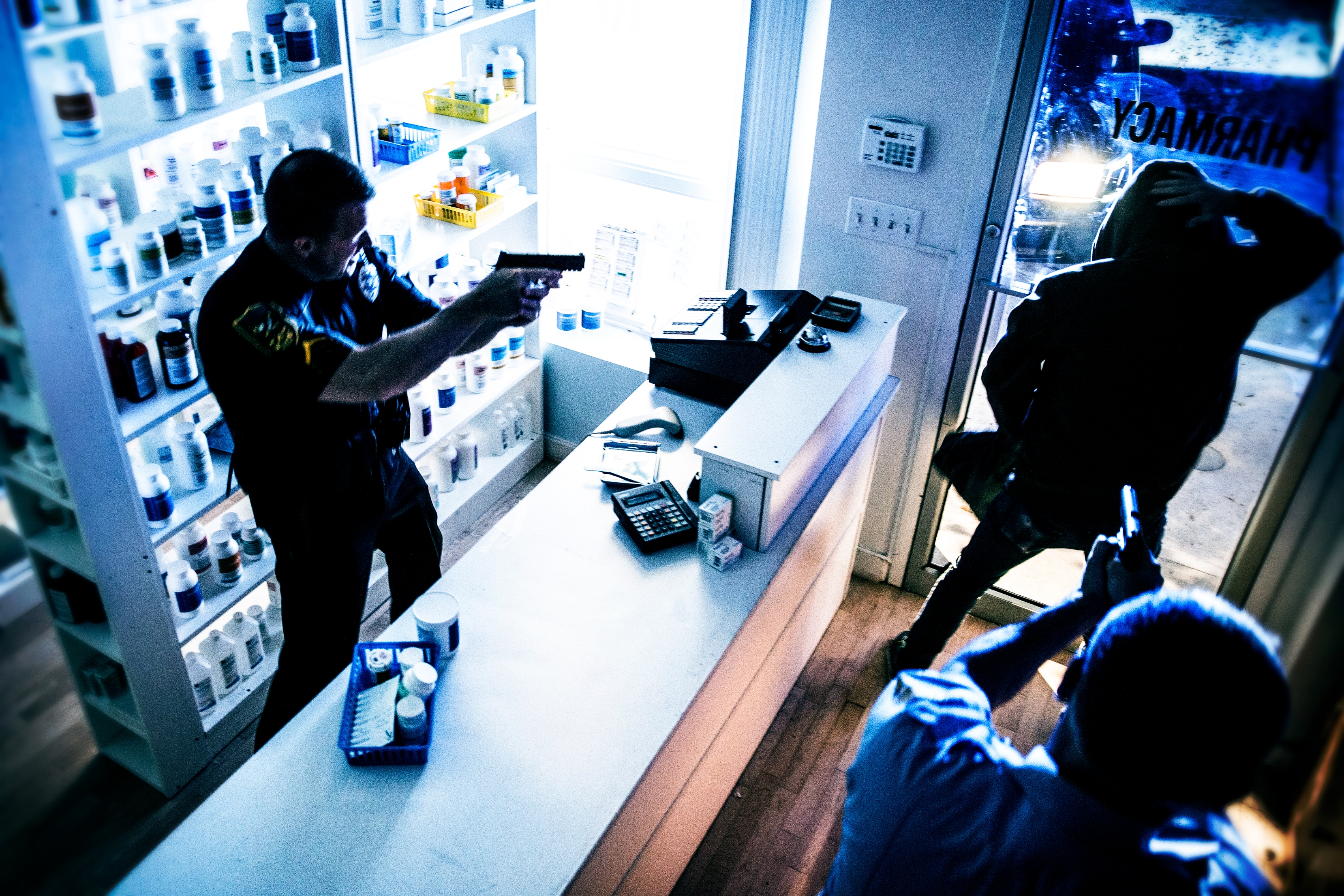 A police officer with a drawn gun confronts a suspect in a pharmacy, with another person in the foreground holding a weapon