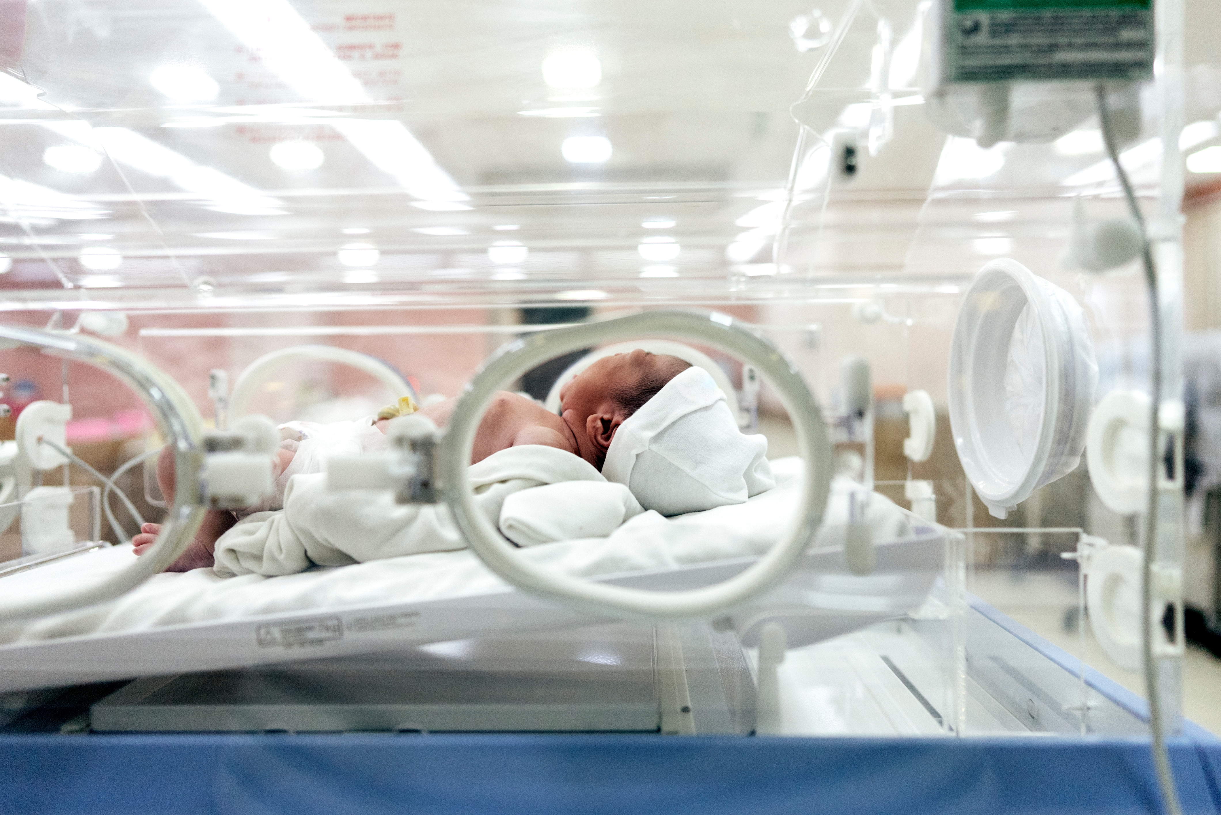 Newborn in an incubator at a hospital, wearing a cap and dressed in white, with medical equipment visible around