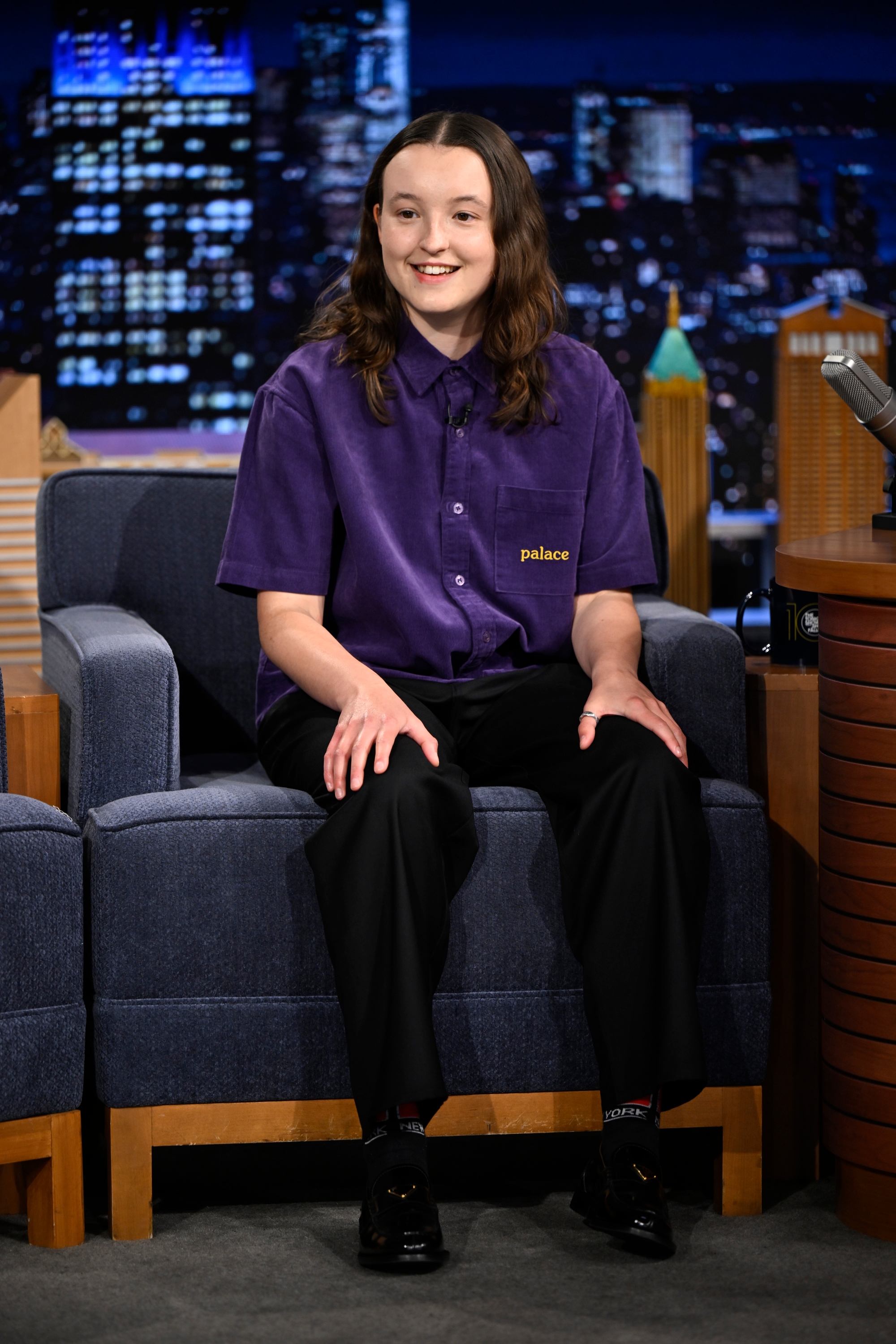 Bella in a talk show studio, wearing a short-sleeve button-up shirt and black pants, smiling while seated and engaging with the audience