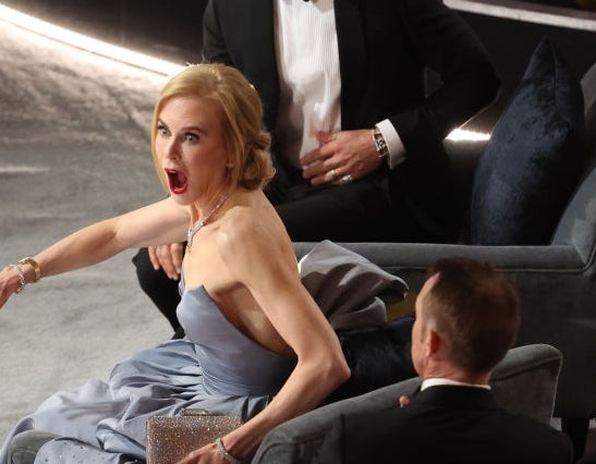 Actor in a strapless gown reacts dramatically while seated at an awards event, with others on chairs nearby