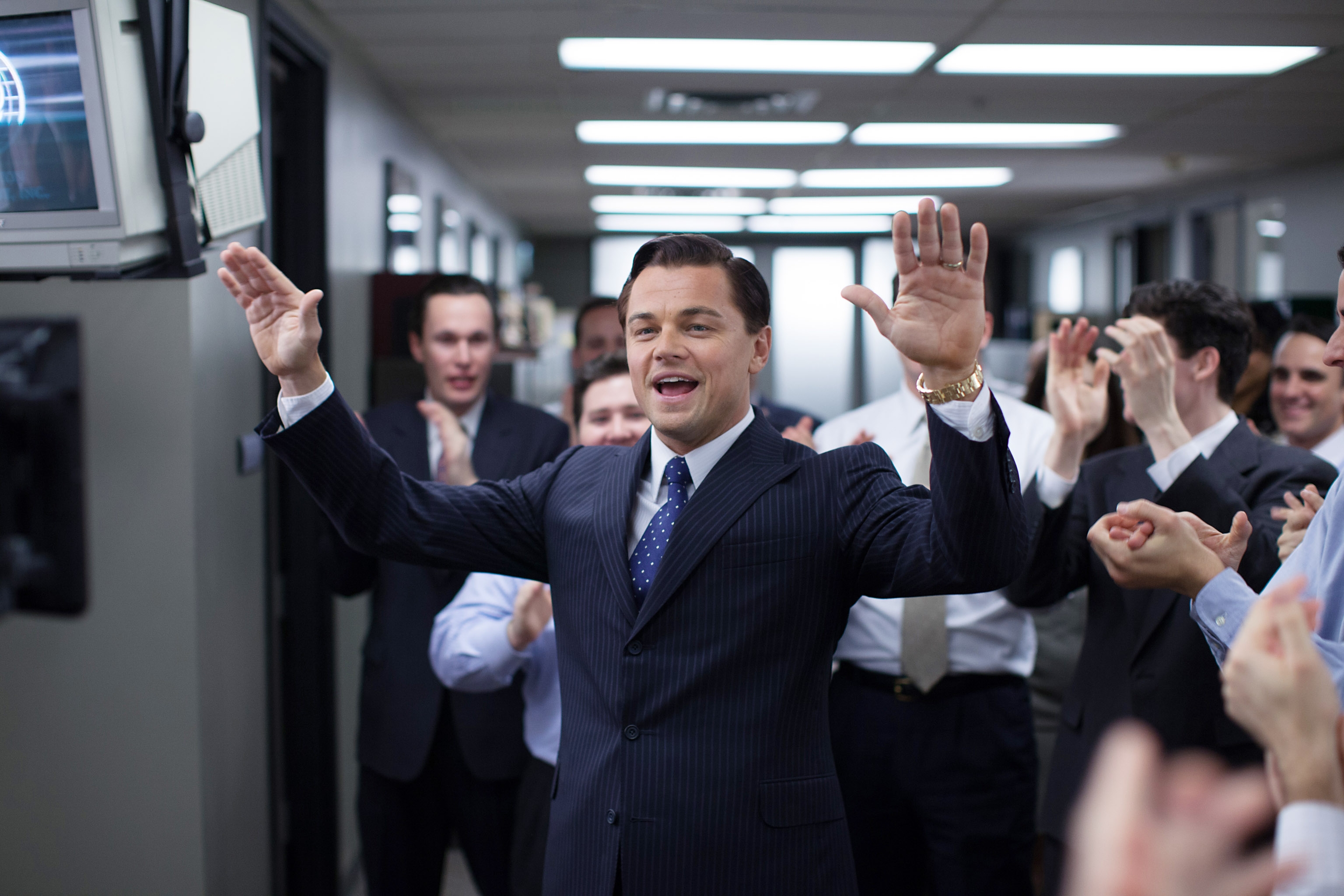 A man in a pinstripe suit stands in an office hallway, surrounded by applauding colleagues