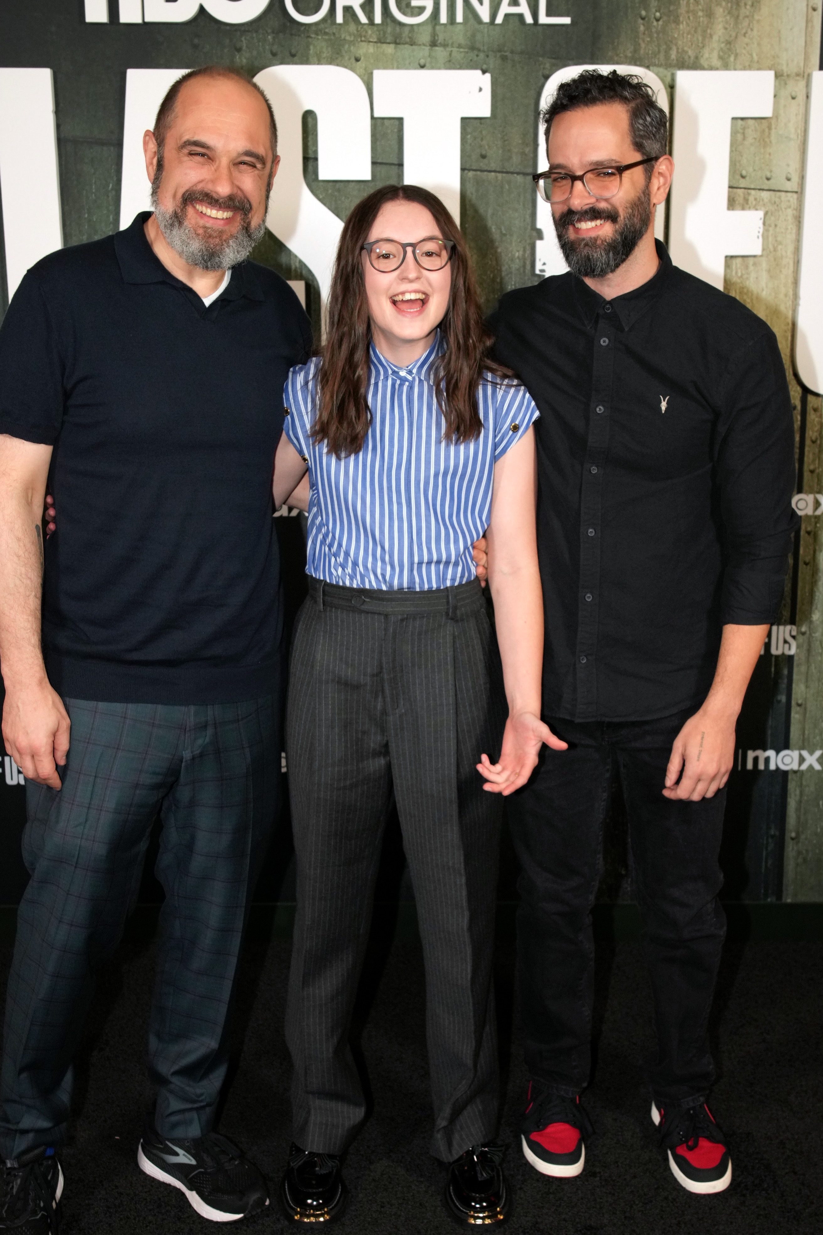 Craig Mazin, Bella Ramsey, and Neil Druckman smiling together on a red carpet