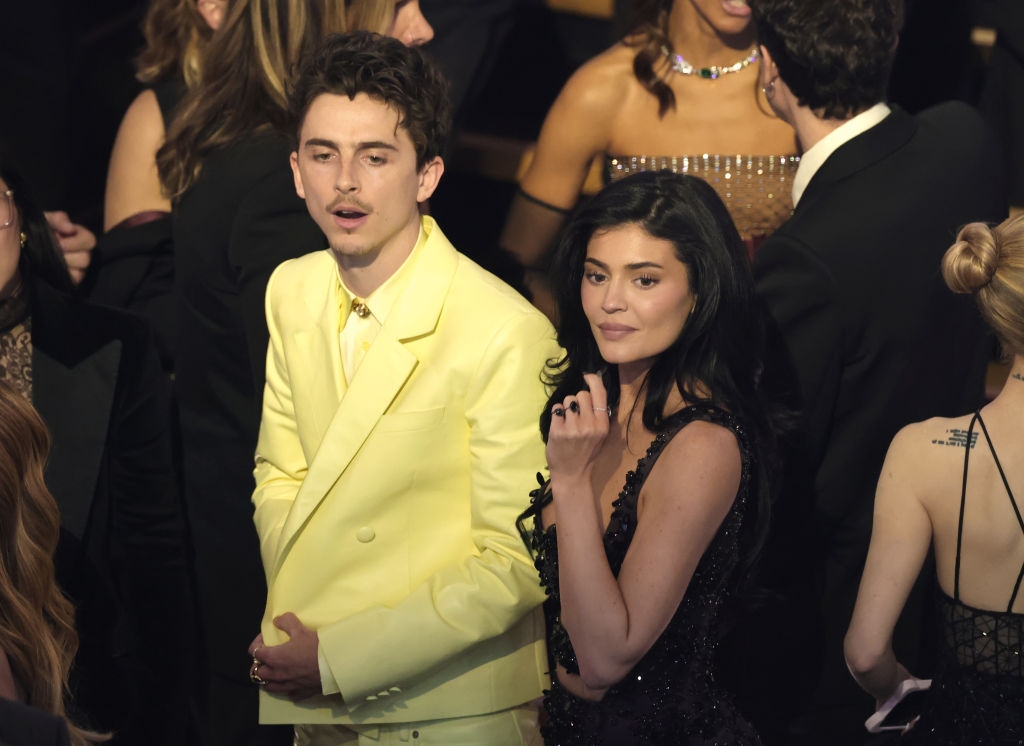 Two people at a formal event. The person on the left wears a yellow suit, and the person on the right wears a black dress. They are in a crowded setting