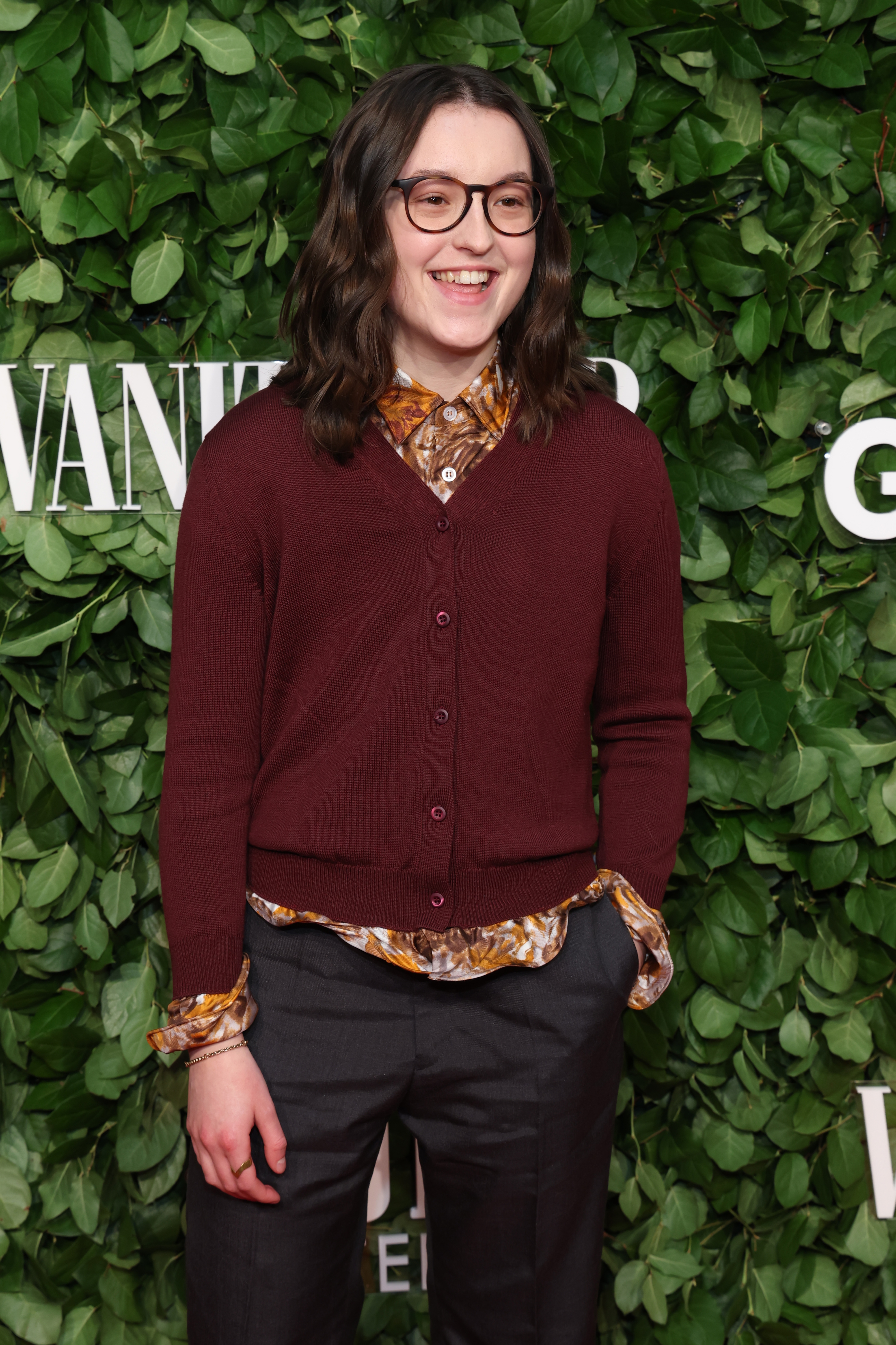 Bella in patterned shirt, maroon cardigan, and glasses stands smiling at a celebrity event with a green leafy backdrop