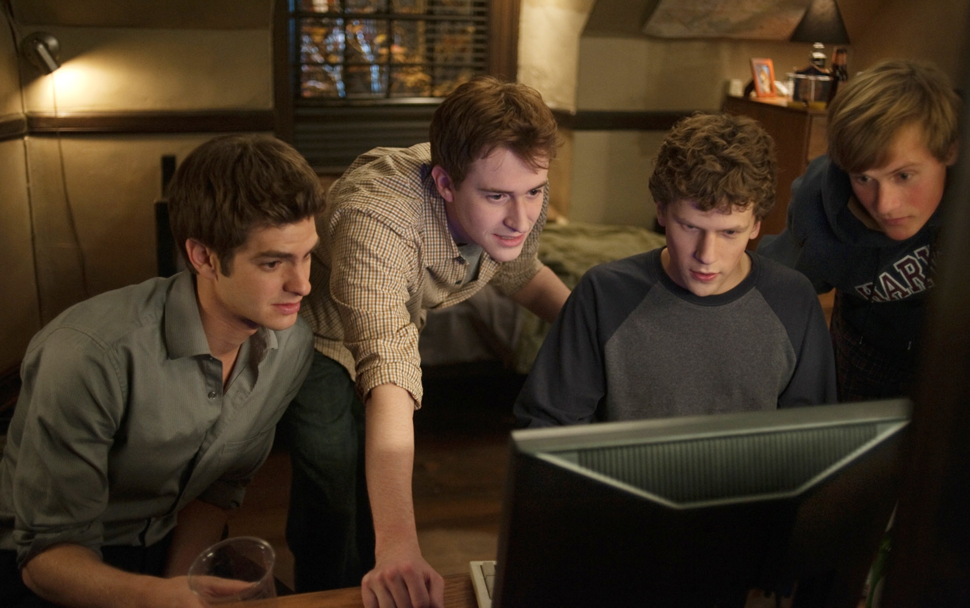 Four young adults huddle around a computer screen, focusing intently on what's displayed. They appear to be collaborating or problem-solving