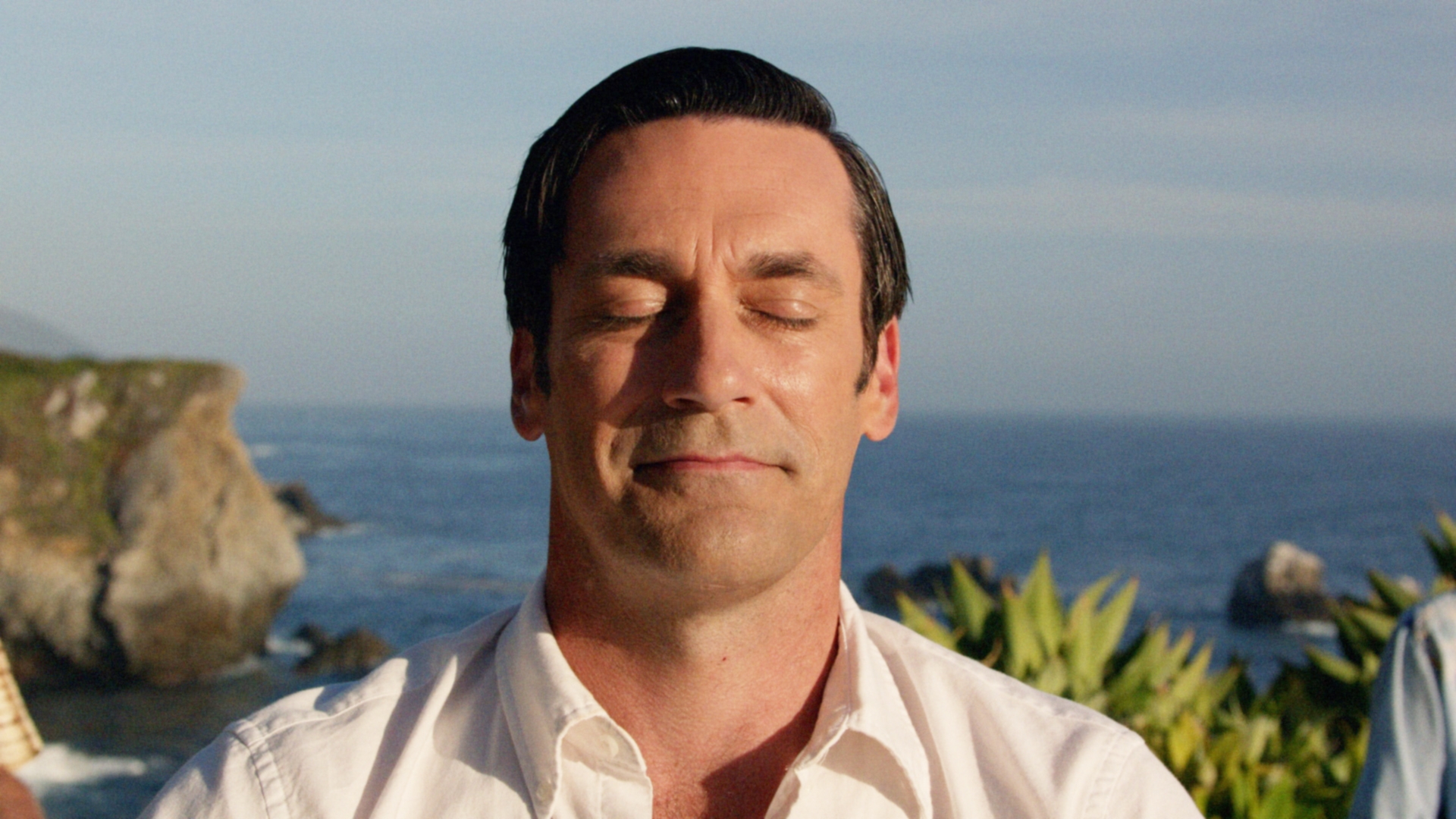 A person with closed eyes smiles gently, standing outdoors by the ocean, wearing a button-up shirt. The background features cliffs and water