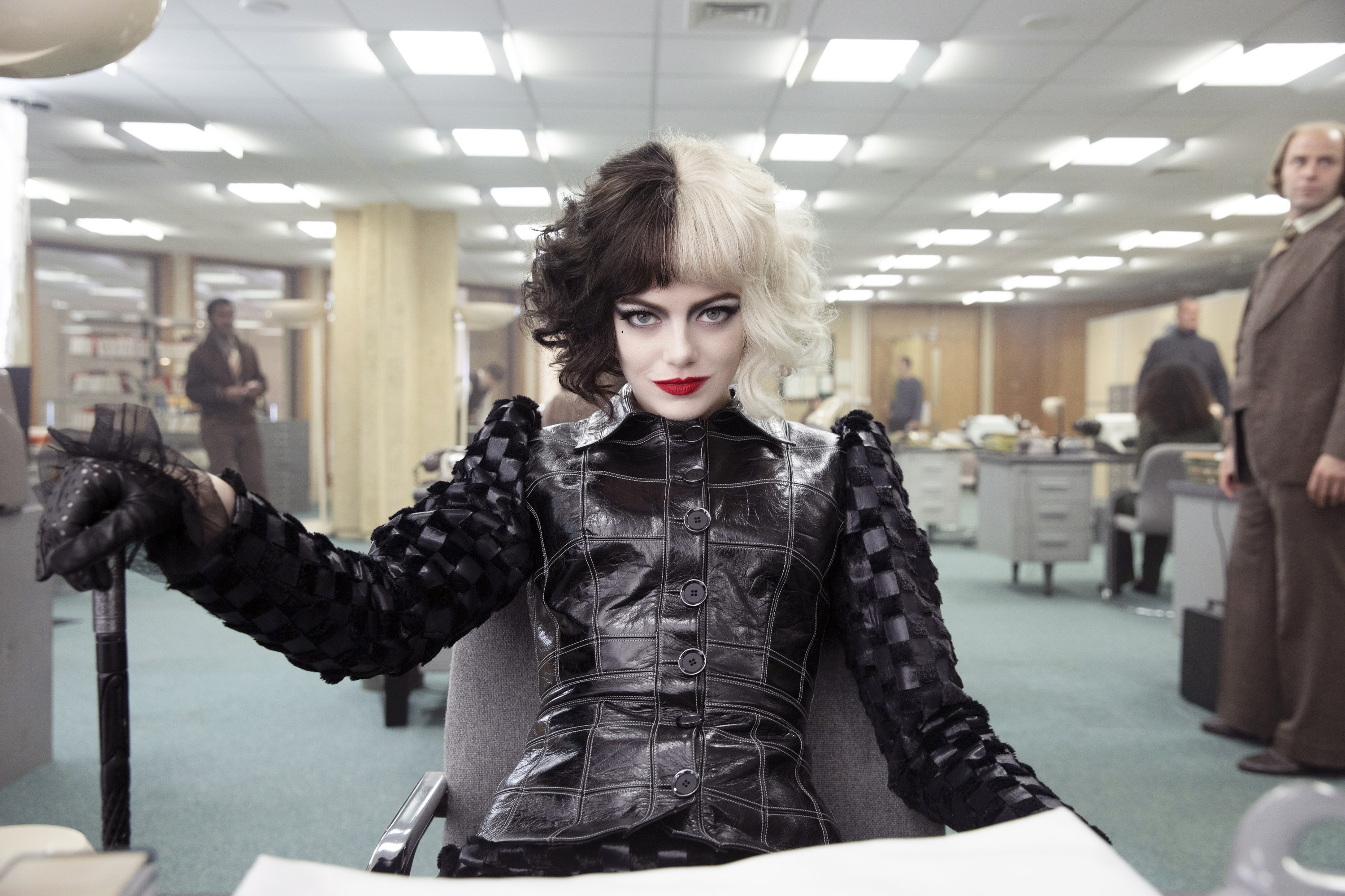 A person in a half-black, half-white wig sits confidently at an office desk, wearing a textured black leather outfit, in a retro-style office