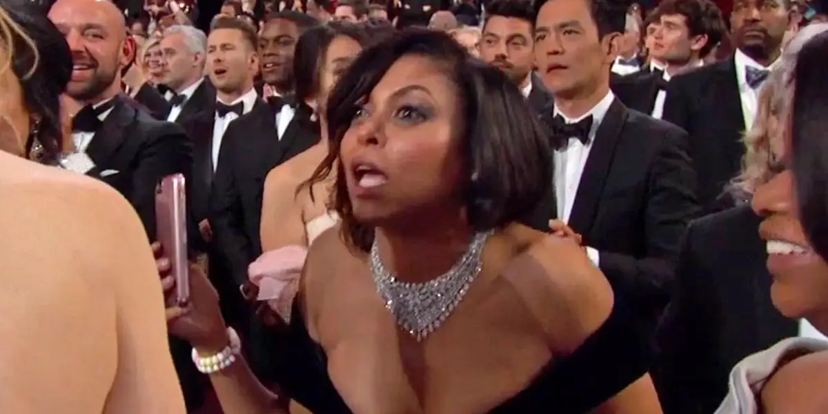 Person in formal attire, adorned with statement jewelry, looking amazed at an event surrounded by an audience in formal wear