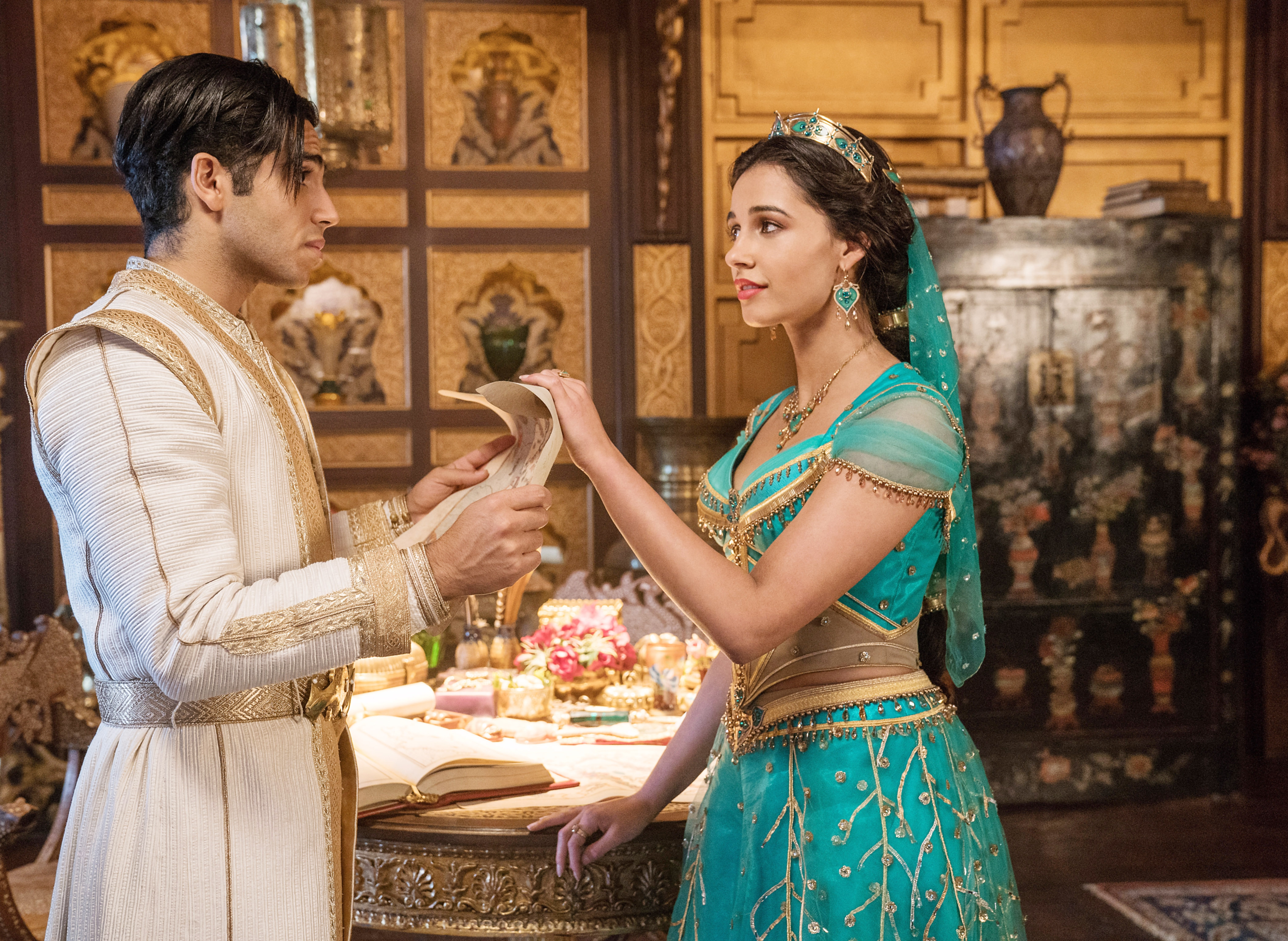 Two actors in a royal setting; one wears a white robe, the other a bejeweled outfit with a headband, interacting over a book