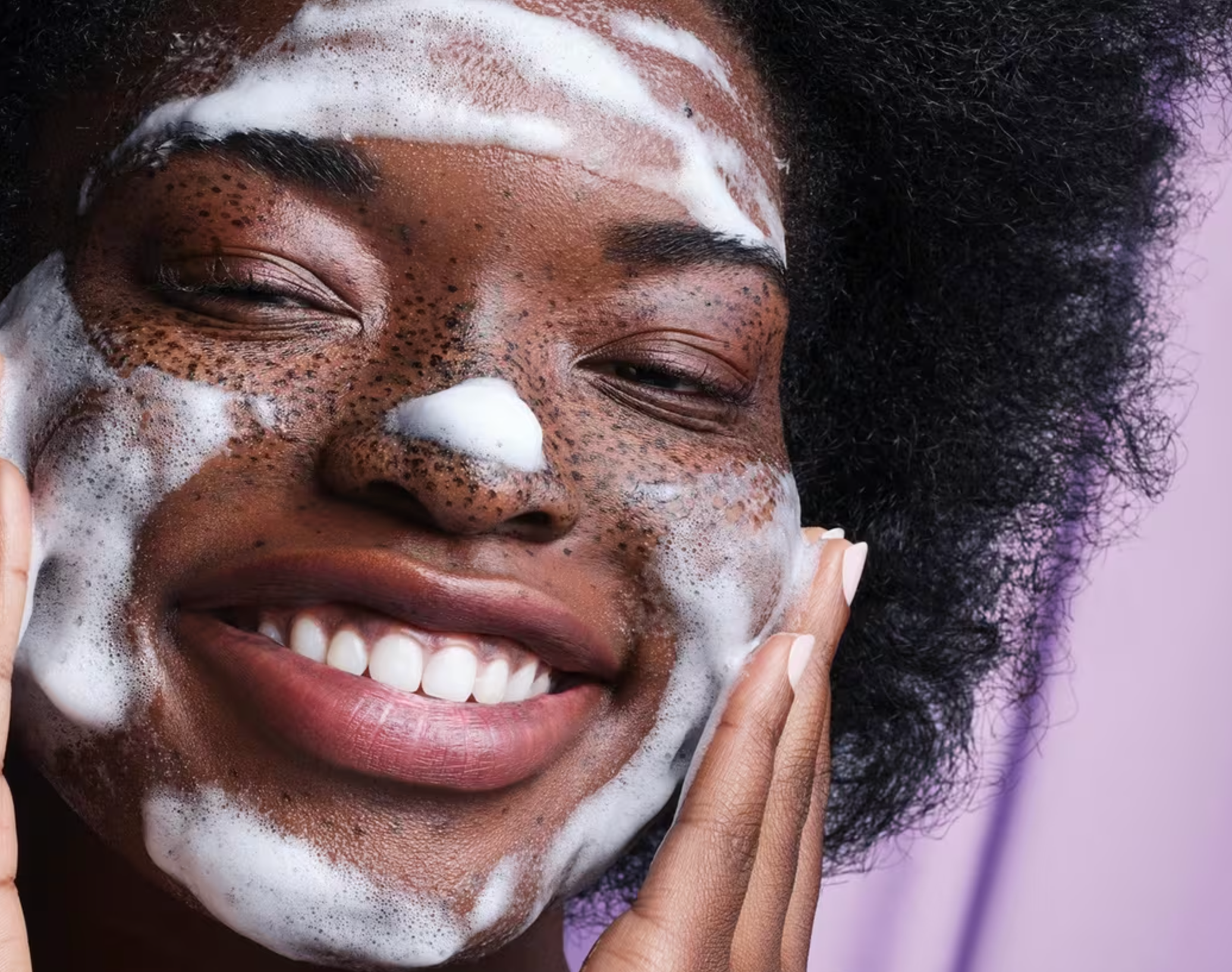 Person smiling portion    applying a foamy facial cleanser, mildly  massaging face, with a purple background
