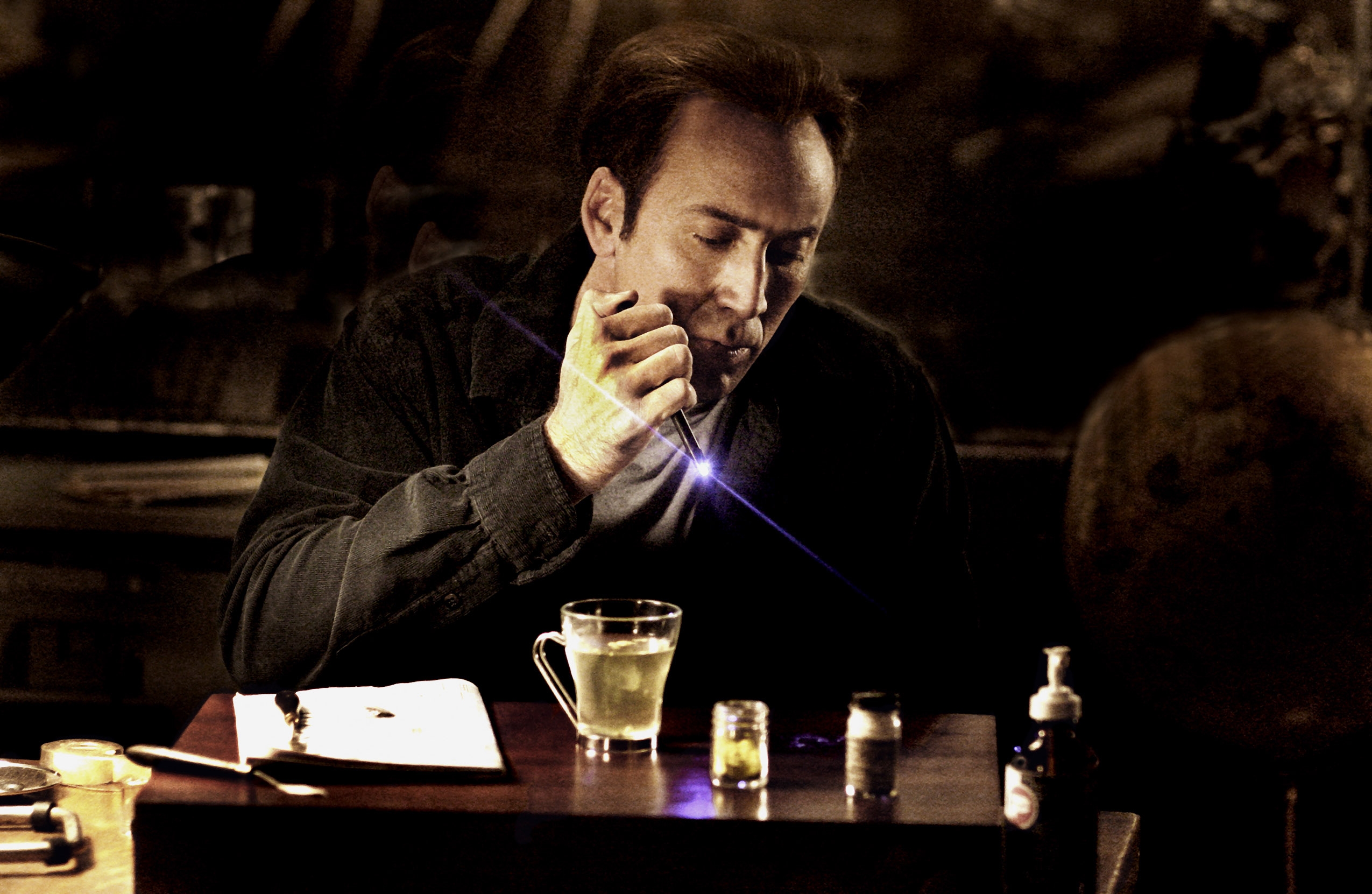 Person examining a glowing object in a dimly lit room, seated at a table with a mug, paper, and bottle nearby