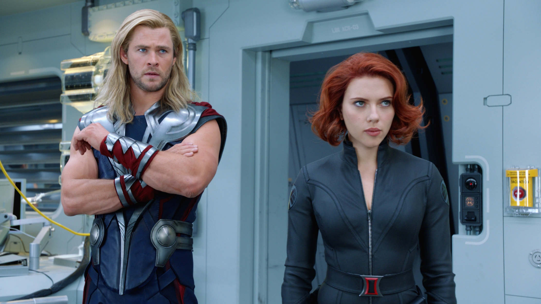 Two actors in superhero costumes are on a set resembling a futuristic laboratory. The man has long blonde hair. The woman wears a zip-up bodysuit