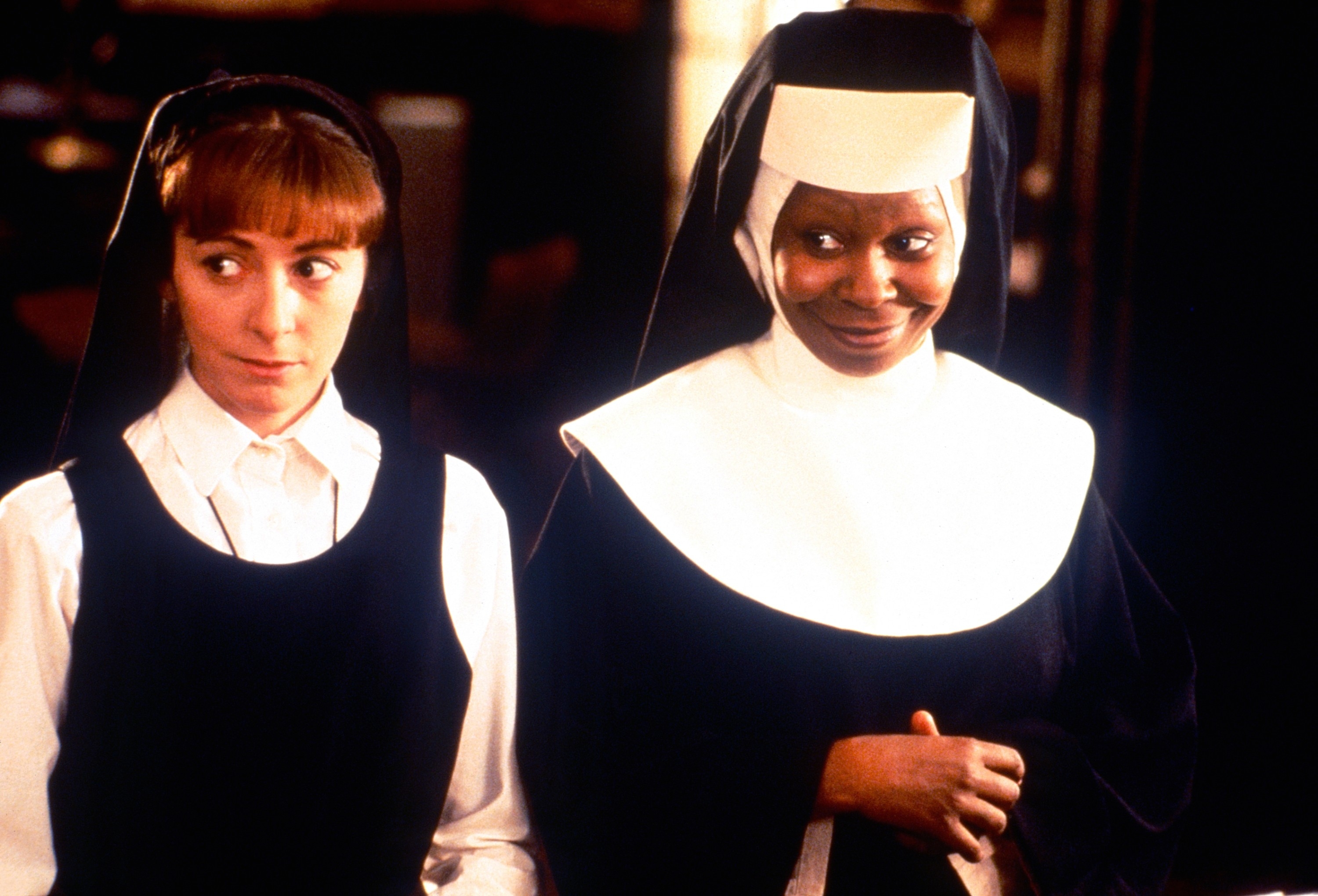 Two women dressed as nuns, one smiling humorously and the other looking unsure, in a scene suggesting a comedic setting