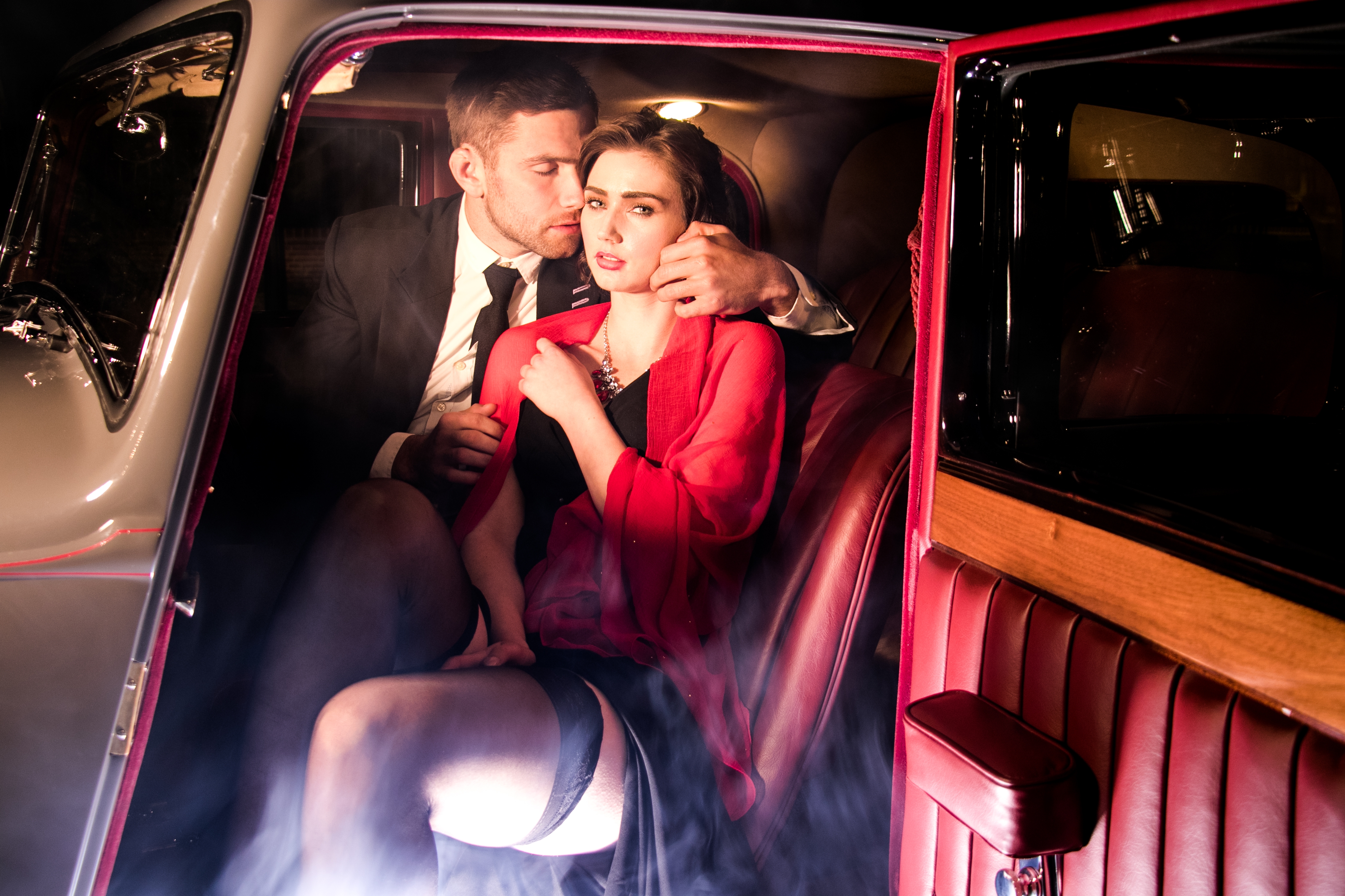 A couple in elegant evening wear sit in a vintage car, with the woman draped in a shawl and the man in a suit, creating an intimate atmosphere