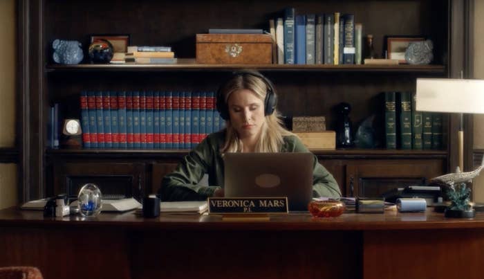 Veronica Mars wearing headphones at a desk with a "Veronica Mars" nameplate, focused on a laptop. Books and office items are on shelves behind