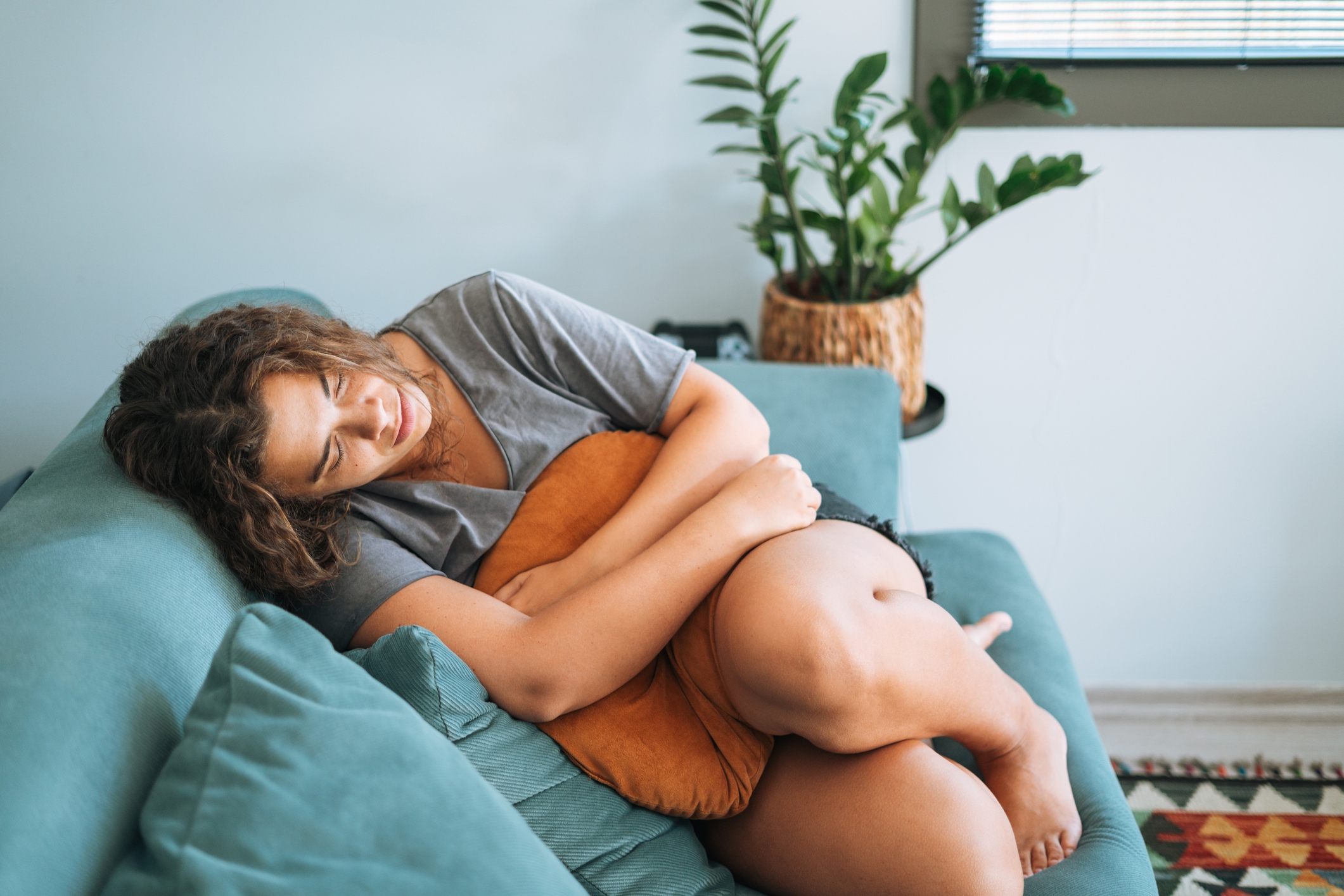 Person lounging on a couch holding a pillow, looking relaxed and thoughtful, with a potted plant in the background