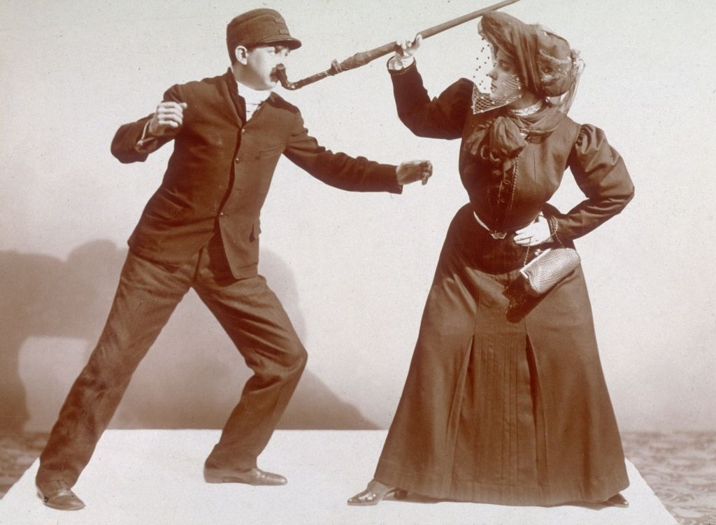 Vintage photo of two people mock-fighting; a woman in a hat and long dress has the handle of an umbrella pushed up against the face of a man in a suit